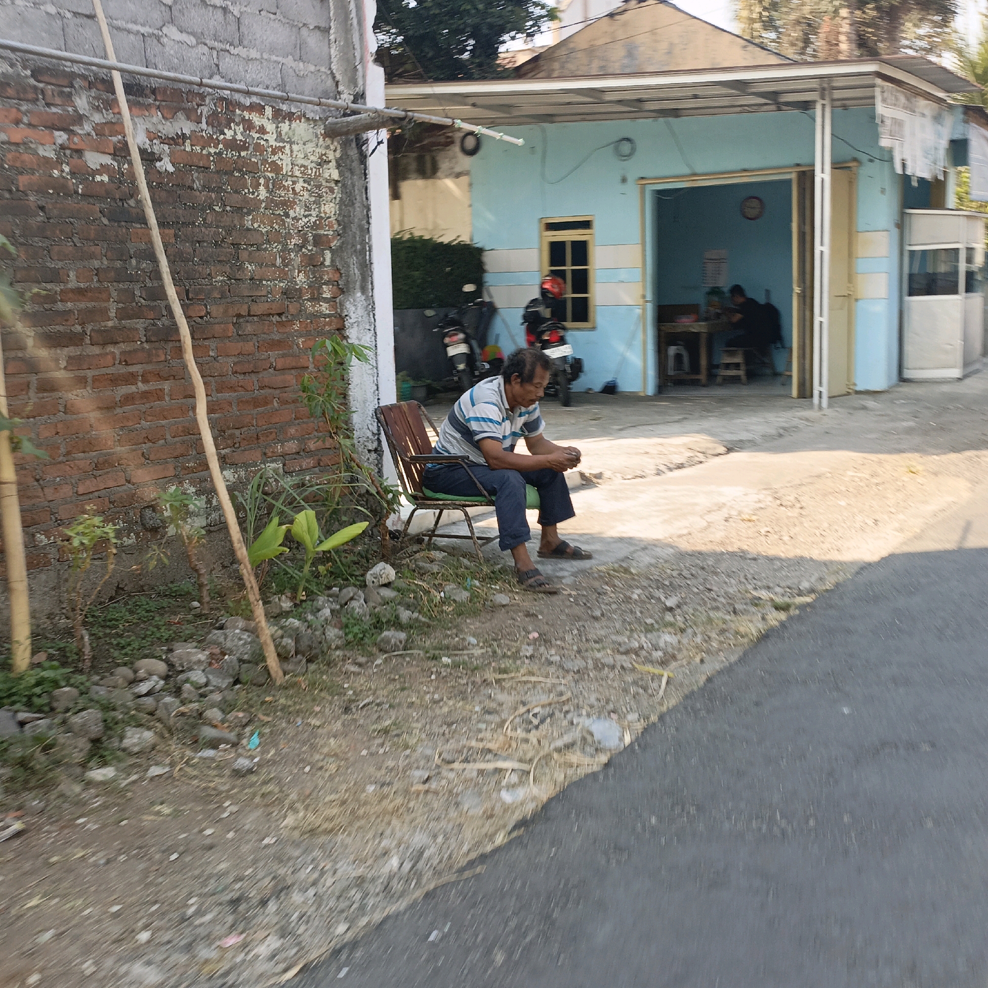 The image shows a man sitting on a chair outside a light blue building with a corrugated metal roof. The building appears to be a small shop or eatery. Inside the building, two people are visible, one sitting at a table and another with their back to the camera. Several motorcycles are parked in front of the building. The man on the chair is wearing a striped t-shirt and dark pants. He is looking down at his hands. To the left of the building is a brick wall, and to the right is a paved road. The ground in front of the building is rough and dusty, with some scattered rocks and vegetation. The scene suggests a casual outdoor setting, likely in a residential or commercial area of Salatiga, Indonesia.