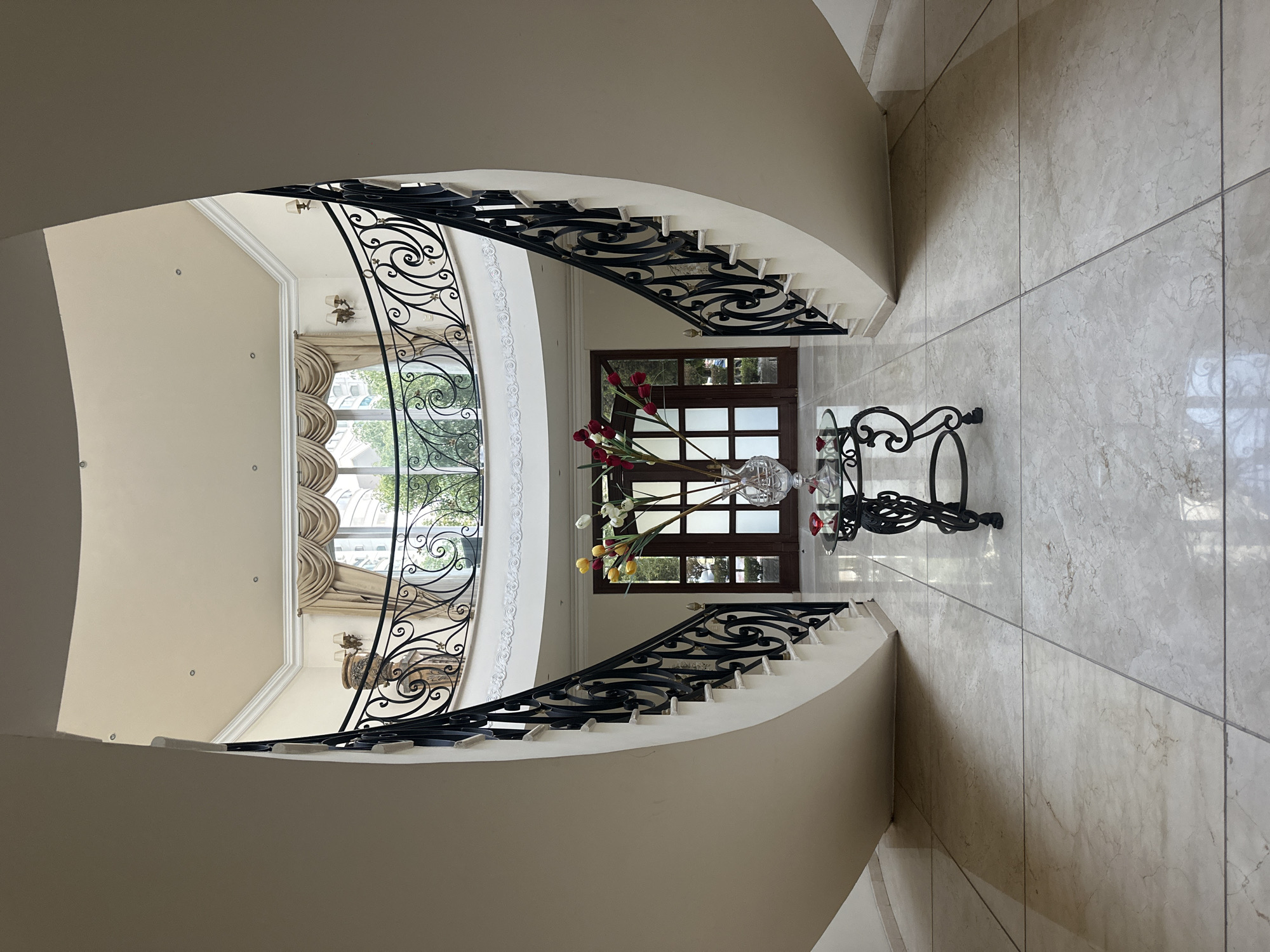 The image depicts the interior of a building, likely a residence, in Jesús del Monte, Mexico. A grand staircase curves around an open space with tiled flooring. A round glass-topped table with a black wrought-iron base is positioned in the center. The table is adorned with a glass vase containing a floral arrangement of red and yellow flowers. In the background, a dark-framed window with sheer curtains offers a glimpse of the exterior. The staircase features a white wall and a black wrought-iron railing with decorative scrollwork. The overall impression is one of luxury and sophistication. No people are visible.