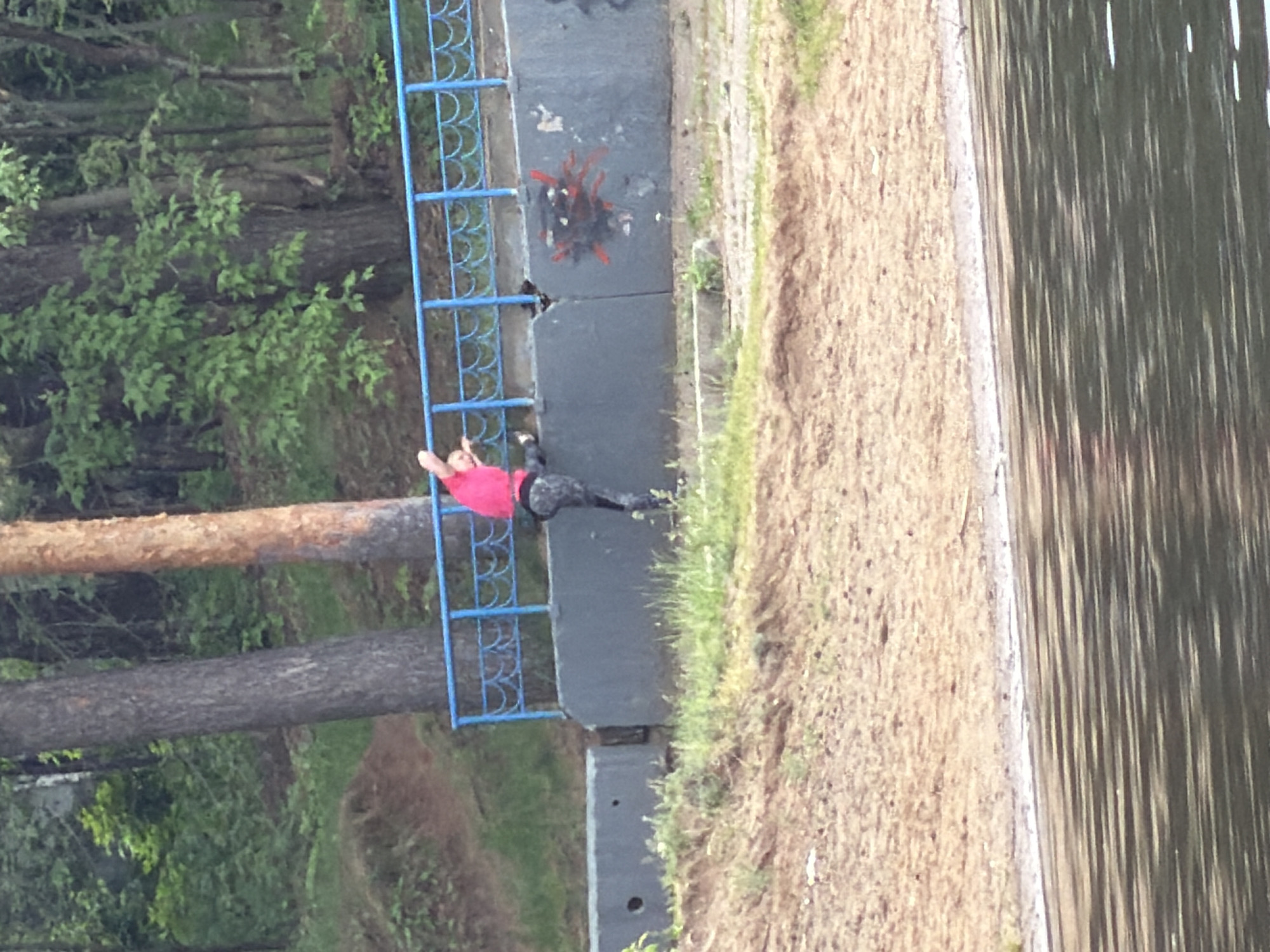 The image depicts a person in a pink shirt and dark pants resting on a blue metal fence along a concrete embankment. The embankment borders a body of water, likely a lake or river, with trees and vegetation in the background. A tree trunk is visible near the fence. The location is described as the city of Chaikovsky, Russia. There are red firework remains on the concrete near the fence.