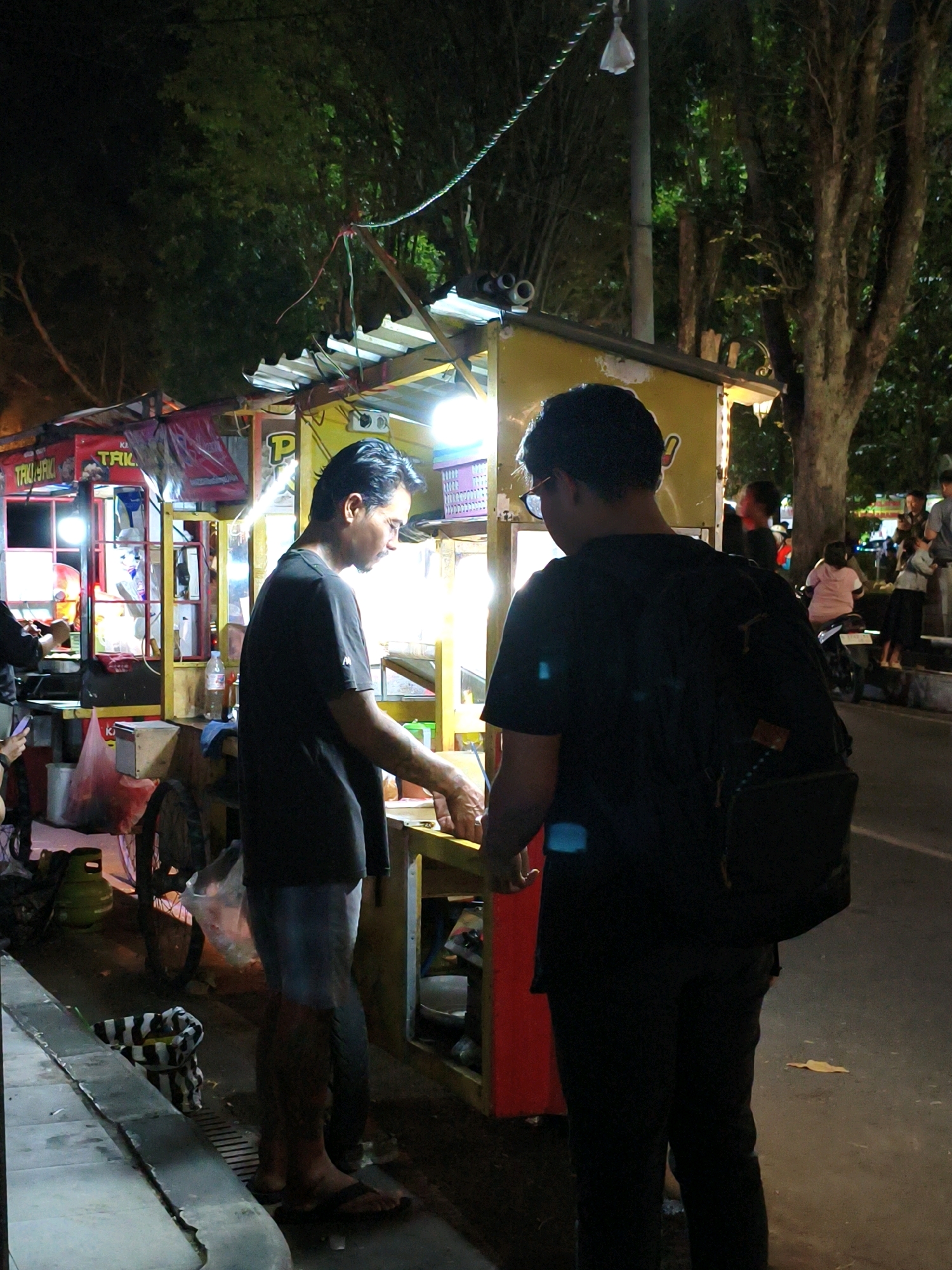 This image depicts a street food vendor and customers at night in Salatiga, Indonesia. The scene is illuminated by bright fluorescent lights from the food stalls, casting a warm glow on the surroundings. The vendor, a man with dark hair and a black t-shirt, is seen preparing food, with his hands engaged in an activity on the counter. He is positioned in front of a yellow food cart adorned with various signs, including one with the letter "P" prominently displayed.
To the right of the vendor, a young man wearing a black t-shirt and carrying a backpack is facing away from the camera, appearing to be a customer or bystander. In the background, other people are visible, including a woman on a motorcycle and several individuals standing around, suggesting a lively street scene. The environment is urban, with trees and a paved road visible. A bicycle with a basket is also present near the food cart. The overall impression is one of a typical evening at a bustling night market or street food hub.