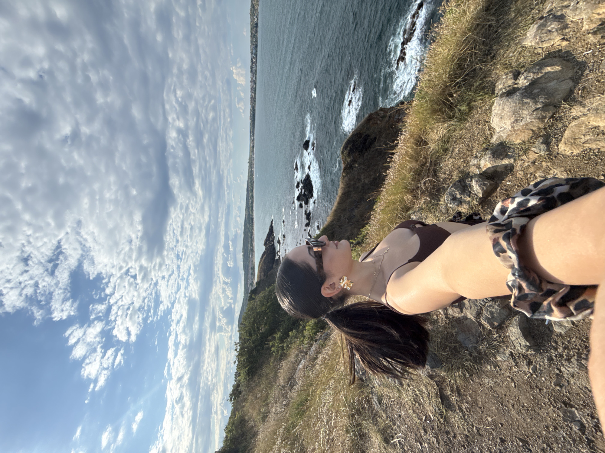 An over-the-shoulder selfie is taken by a woman with dark hair in a ponytail, wearing sunglasses and a brown bikini top. She is standing on a rocky, grassy cliff overlooking the ocean. The water is a deep blue with white waves crashing against rocky outcrops. The sky is partly cloudy with dramatic cloud formations. In the distance, a town or city is visible along the coastline. The scene appears to be a scenic overlook.