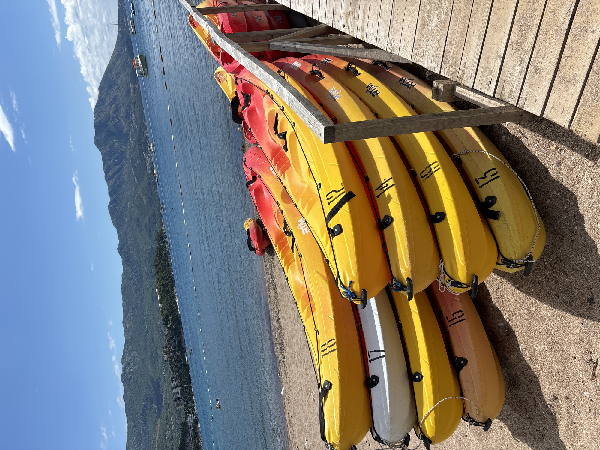 The image captures a sunny day at a beach in Budva, Montenegro, with a focus on a stack of kayaks on the sand next to a wooden pier. The kayaks are predominantly yellow, red, and orange, with some also white, and are neatly piled on top of each other. Numbers are visible on some of the kayaks, including '18', '17', '15', and '13'. In the background, the turquoise waters of the Adriatic Sea stretch out, with a few boats and yachts visible on the horizon. Lush green mountains form a dramatic backdrop to the scene, with some buildings nestled on the slopes. A person is seen swimming or wading in the water to the left of the frame. The wooden pier runs along the right side of the image, suggesting a common area for beach activities.