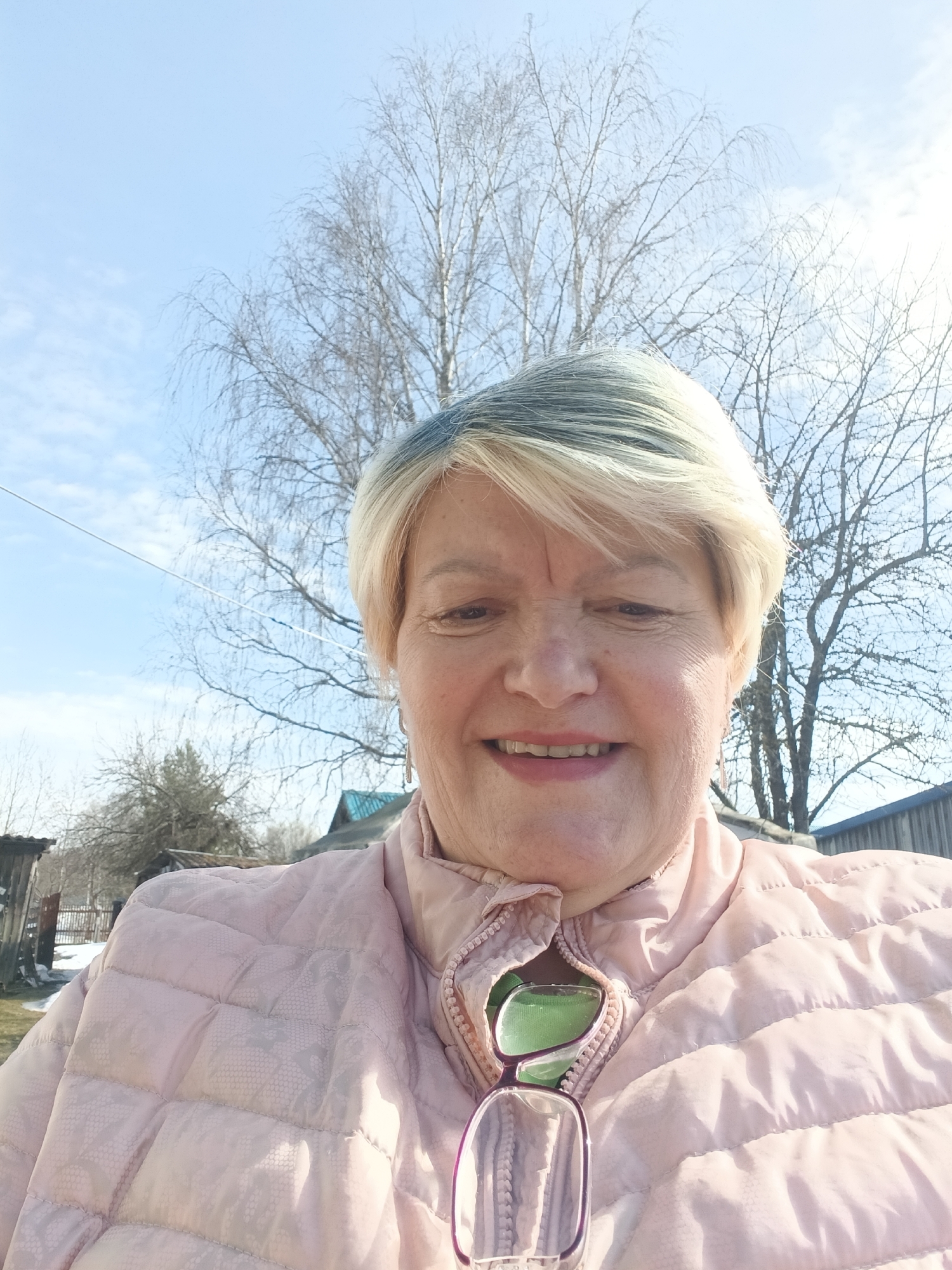 An adult woman is smiling in an outdoor selfie on a sunny day in Красный Маяк, Russia. She has short blonde hair with grey roots, and wears a light pink quilted jacket with glasses tucked into the neckline. The background features bare trees silhouetted against a pale blue sky with scattered clouds. A fence and a wooden building are visible in the background.