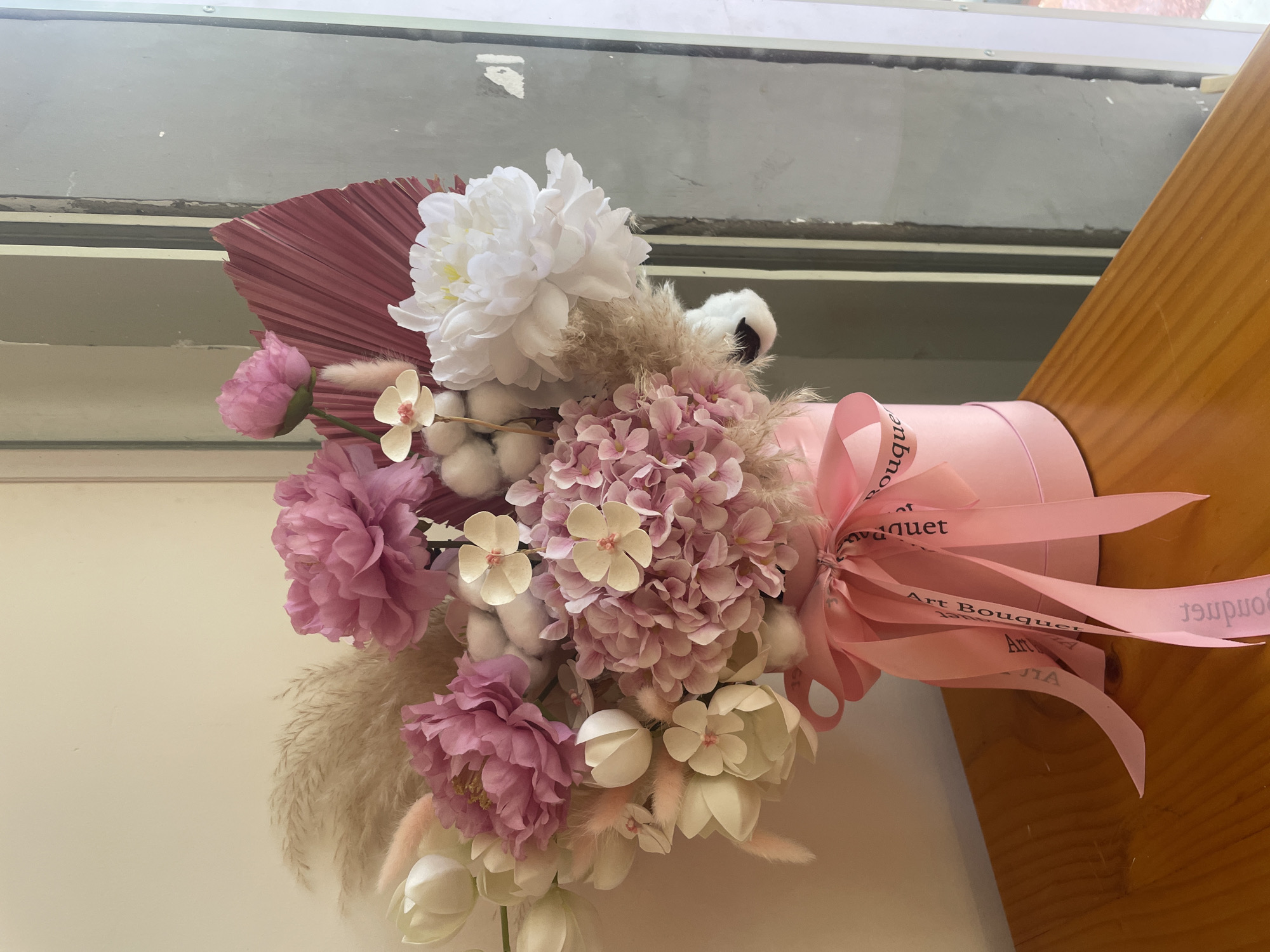 This media file captures a close-up of a beautifully arranged artificial floral bouquet, set indoors. The bouquet is presented in a cylindrical pink box or vase and adorned with a pink satin ribbon.
The arrangement features a variety of flowers and decorative elements in soft pastel tones. Large white peonies are prominent, alongside smaller pink and mauve peonies. Clusters of light pink hydrangeas form a central part of the bouquet, complemented by cream-colored, orchid-like flowers and white tulip-shaped blossoms. Interspersed throughout are decorative fillers such as white cotton balls, light brown feathery pampas grass-like plumes, and smaller pink fluffy elements. A striking dark pink or burgundy dried palm fan adds a textural and structural accent to the left side of the arrangement.
A pink satin ribbon is tied around the base of the pink box, featuring the text "Art Bouquet" repeated in a darker shade, visible in several places, though sometimes partially obscured or reversed.
The background reveals a window with glass panes, showing reflections and some wear, suggesting an outdoor scene beyond, consistent with the provided location context of Bac Nha Trang Ward, Vietnam. To the right, a wooden surface, likely a table or countertop, holds the bouquet. The lighting is soft and diffused, indicating natural daylight from an indoor setting. The overall aesthetic is one of delicate beauty and elegance.