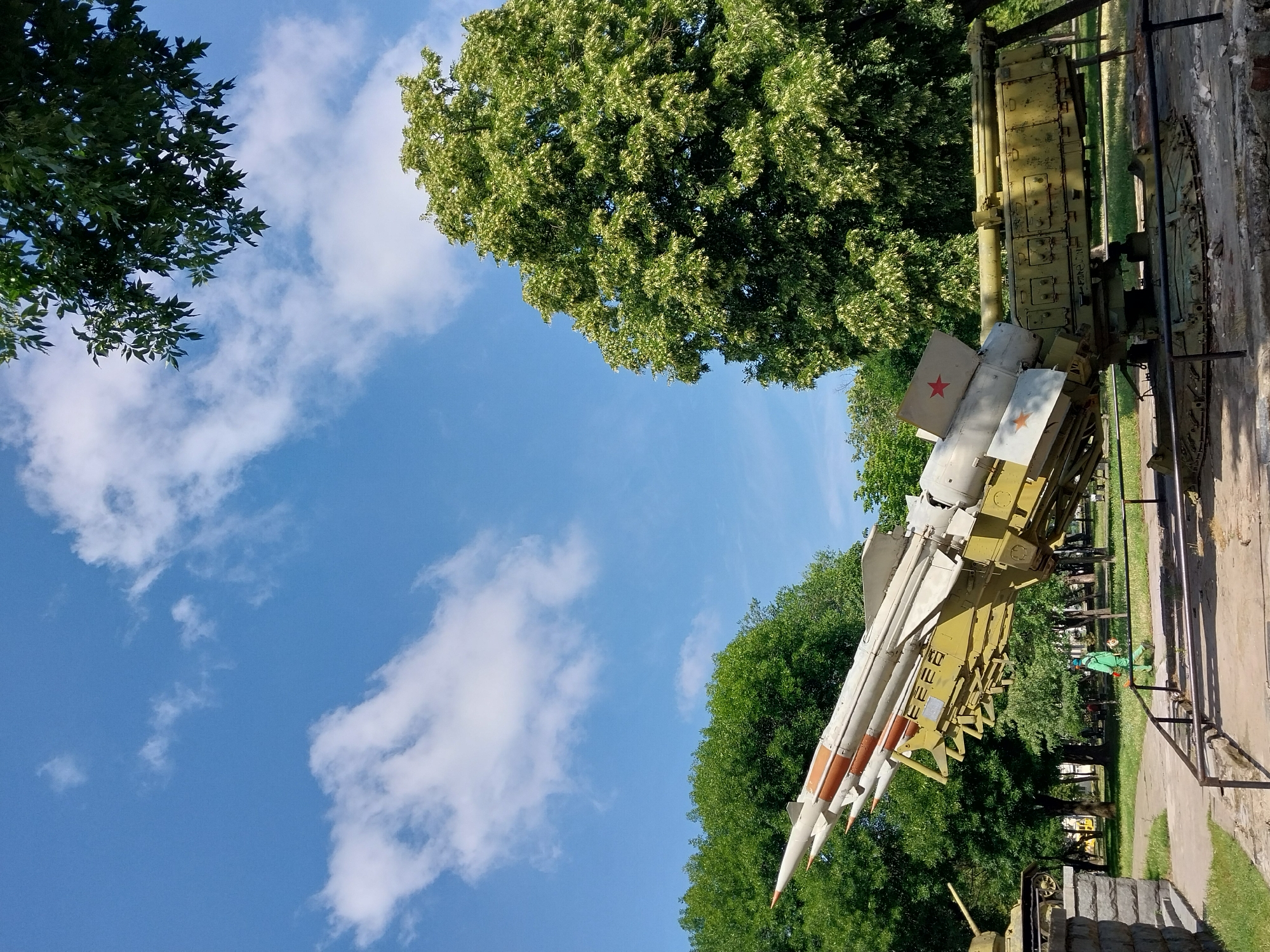 In Dnipro, Ukraine, at the National Museum of Ukrainian Aerospace Technology, a large missile launcher with three missiles is on display. The launcher is painted olive green and is positioned outdoors, angled upwards. The missiles are white with orange and grey accents, and two of them bear red stars. To the left of the launcher, a part of a tank is visible, also on display. A person wearing a green uniform with a blue helmet is walking near the launcher. The background is filled with lush green trees, and the sky is blue with scattered white clouds. The museum grounds appear to be paved in some areas and grassy in others, with stone and metal barriers surrounding the exhibits.