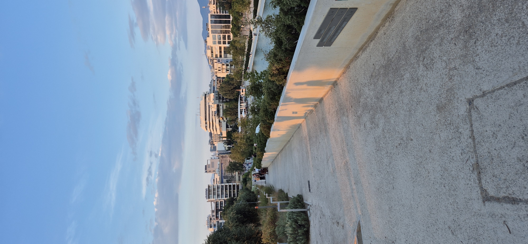 The image depicts a pathway in Kallithea, Greece, likely at the Stavros Niarchos Foundation Cultural Center, given the architectural style and waterfront view. The pathway is paved with concrete and slopes slightly downwards. On the right, a low, cream-colored wall runs alongside the pathway, casting long shadows from figures, suggesting late afternoon or early evening light. Two dark metal plaques are affixed to this wall. Beyond the wall, lush green bushes and trees line the edge of a body of water, likely part of the Stavros Niarchos Park. Across the water, a modern cityscape is visible, characterized by multi-story buildings with clean lines and large windows. Some of these buildings feature distinctive curved designs. A few people are walking along the pathway in the distance, and a small group is gathered near the start of the pathway. The sky is a clear blue with scattered, wispy clouds.