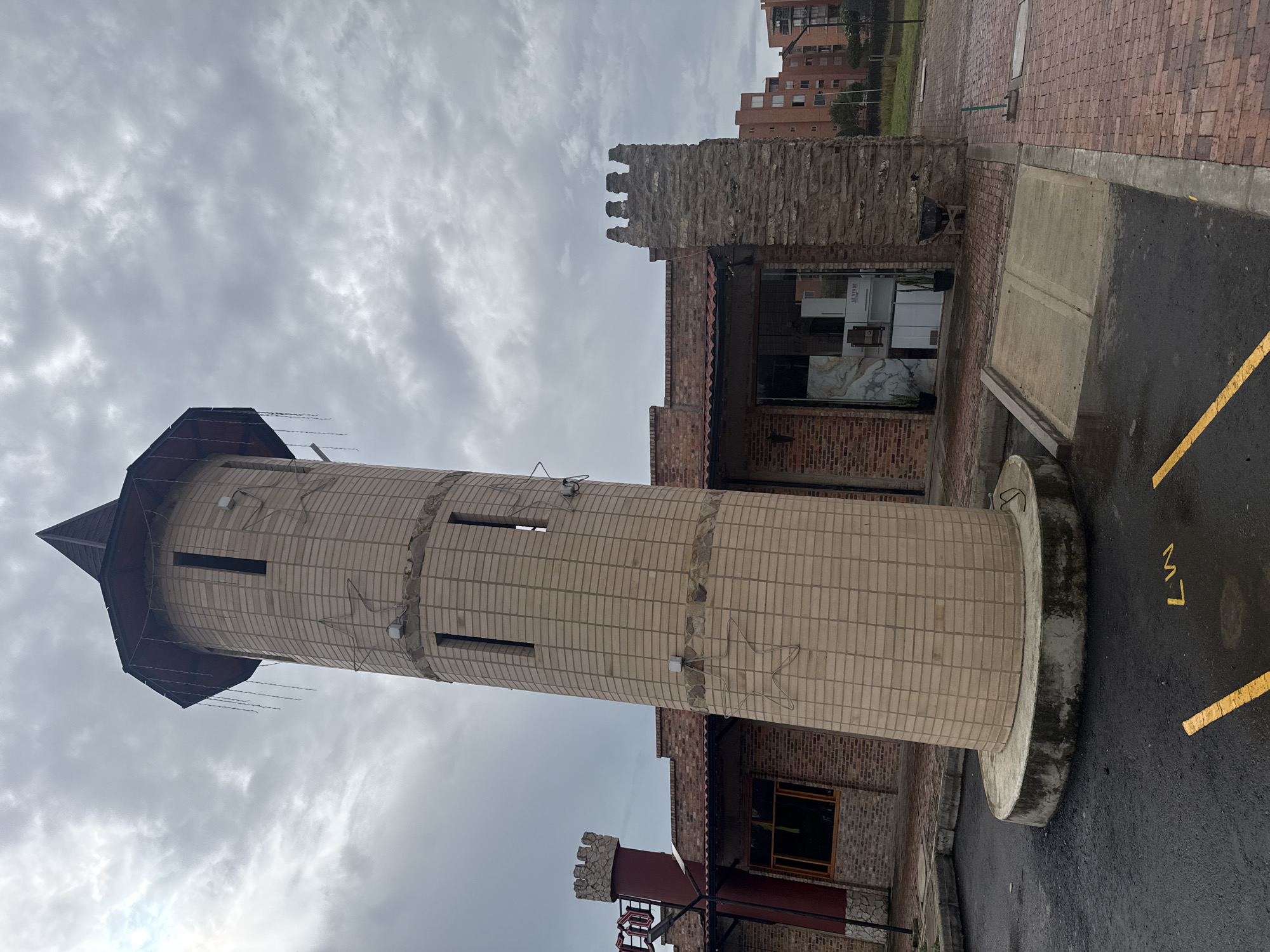 This image, captured in Madrid, Colombia, depicts a castle-themed commercial building under an overcast sky. The prominent feature is a tall, cylindrical tower constructed from light-colored bricks, featuring a dark conical roof with hanging decorative elements resembling string lights. The tower has multiple narrow, vertical window openings and two star-shaped light frames affixed to its exterior. It rests on a circular concrete base, showing signs of weathering and dark stains.
Attached to the tower is a single-story building primarily constructed from reddish-brown bricks with a red tiled roof. The building incorporates architectural elements like battlements on its roofline and a smaller, red-colored, turret-like structure with battlements on the left. The right side of the building features a section clad in rough-cut stone, which also includes battlements. A large glass storefront window on the right displays what appears to be large marble slabs and white kitchen appliances, suggesting a showroom or retail space. Two potted plants flank this entrance. Another smaller window with a brown frame is visible on the left. A small black wheelbarrow is positioned near the stone-clad wall on the right.
The foreground consists of a dark asphalt parking area with yellow painted lines and the characters "LW M7". The ground appears wet, suggesting recent precipitation. A concrete ramp leads from the asphalt to a brick-paved sidewalk or patio area that extends to the building's entrance. In the distant background, several multi-story residential buildings constructed of reddish-brown materials are visible under the cloudy sky. No individuals are present in the scene.