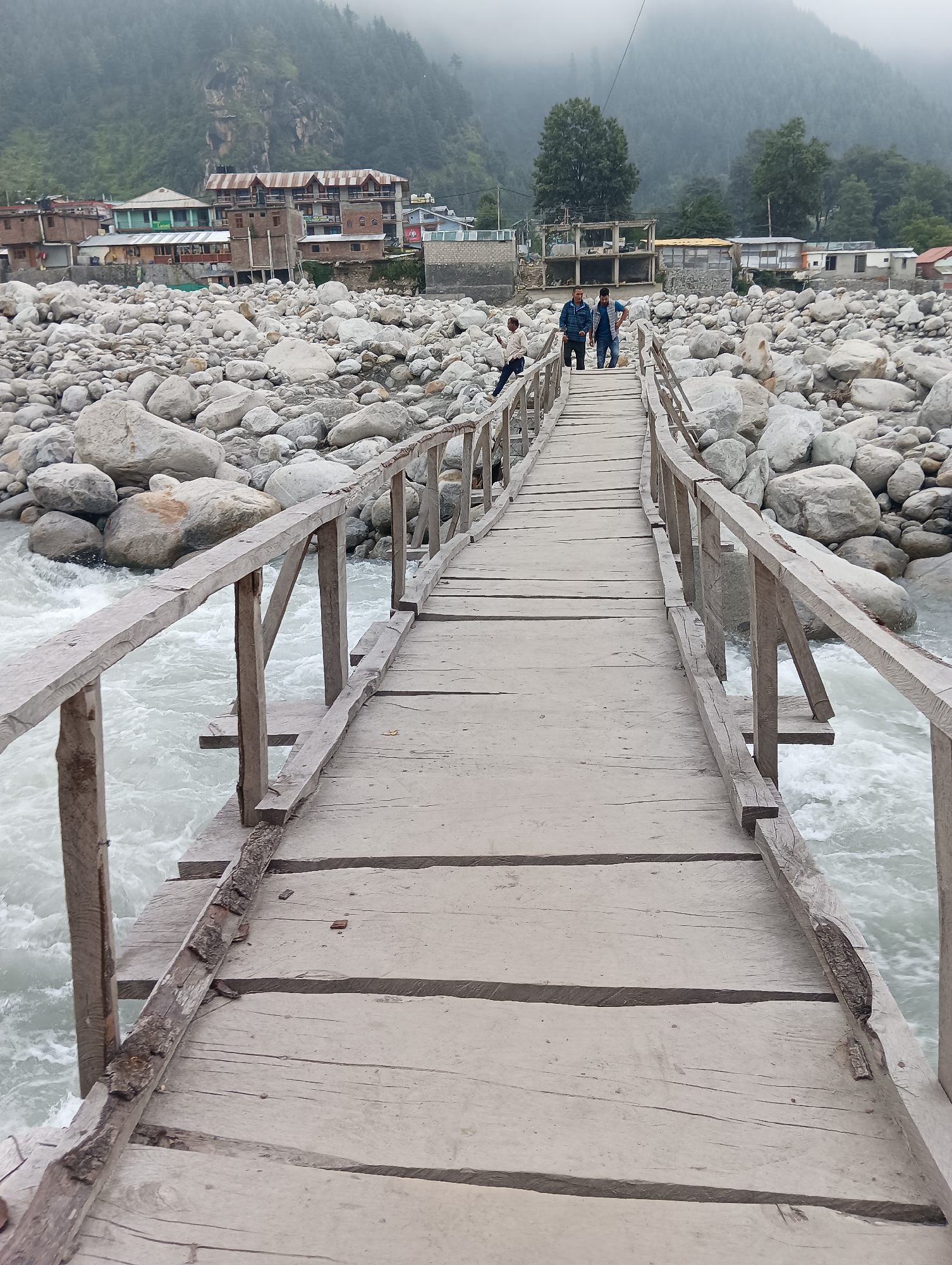 A wooden footbridge spans a rocky riverbed in Bahang, India. The bridge, constructed with weathered planks and railings, shows signs of age and wear. Three individuals are present on the bridge. Two men in casual attire, one wearing a blue jacket and the other a dark shirt, walk towards the viewer. A third person, partially visible, is seated on the left railing, looking away. The river below flows rapidly with white water, surrounded by a multitude of large, smooth stones. In the background, the town of Bahang is visible, characterized by a collection of buildings with varied architectural styles, including multi-story structures with corrugated metal roofs and some under construction. The scene is set against a backdrop of mist-shrouded mountains and dense coniferous trees, suggesting a natural and somewhat remote environment.