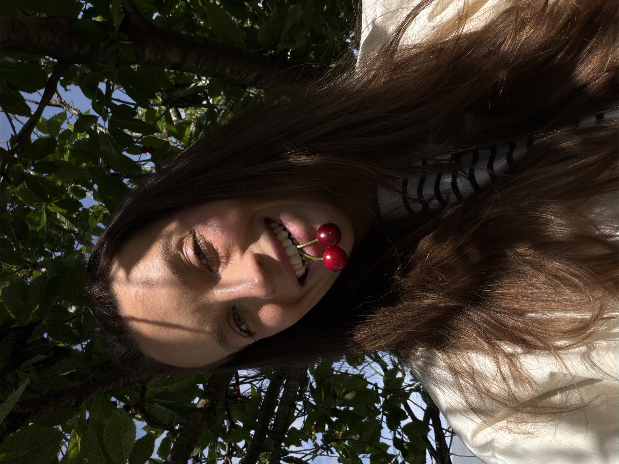 A woman is holding two cherries in her mouth, lying on her back, looking up at the camera. She is positioned under a tree with green leaves. The sky is visible through the leaves. The location is Mojanovići, Montenegro.