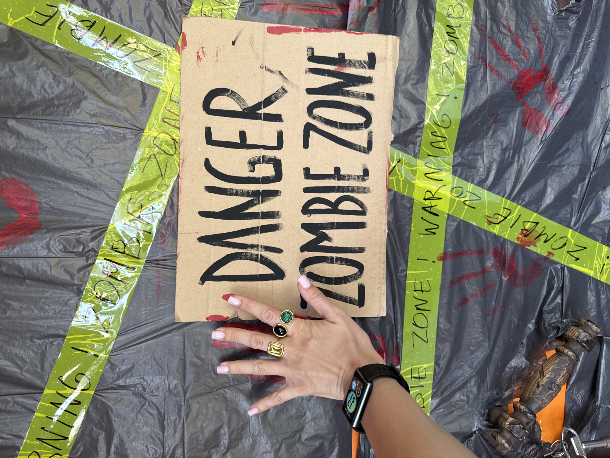 The image depicts a close-up view of a hand adorned with rings and a smartwatch, positioned in front of a cardboard sign that reads "DANGER ZOMBIE ZONE." The sign is affixed to a black plastic sheeting, which is further decorated with red handprints and yellow caution tape. The caution tape also has text, portions of which are visible and appear to read "WARNING" and "ZOMBIE." A wooden object, possibly a prop, and keys are visible in the lower left corner of the frame. The background is dark and textured, suggesting an indoor or shaded outdoor setting. There is no information in the image to suggest a location in Sochi, Russia.