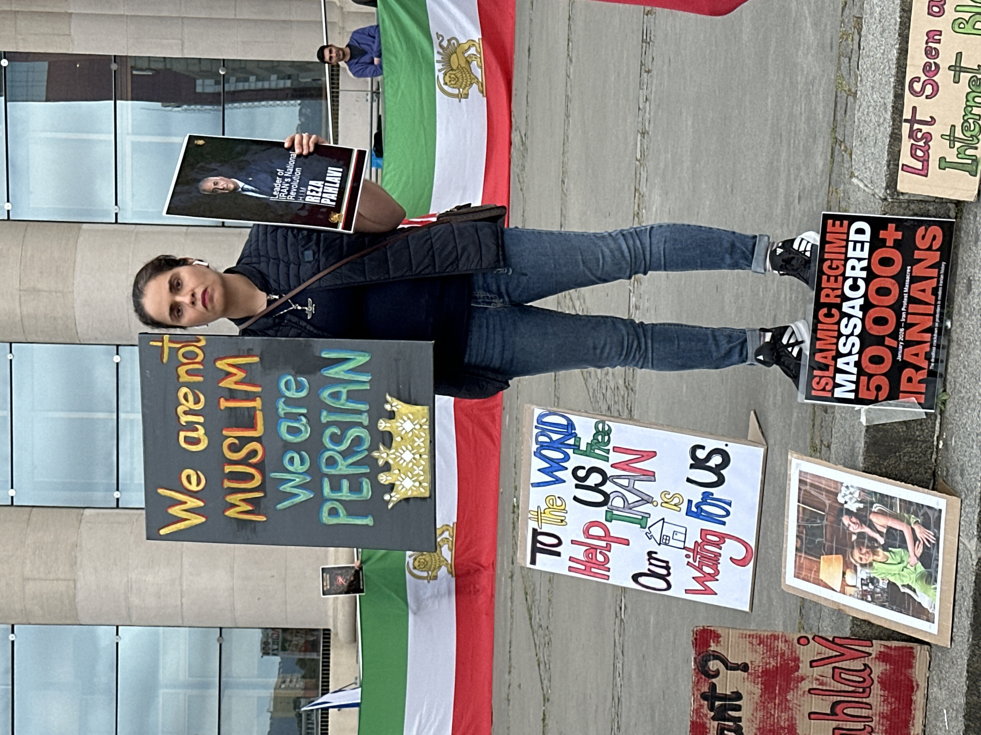A demonstrator stands on a paved surface holding a sign reading "We are not Muslim, We are Persian" next to an Iranian flag. Additional signs and photographs lie on the ground. One sign says "Leader of Iran's National Revolution, H.I.M. Reza Pahlavi" with a portrait. Another says, "To the world help us free Iran. Our is waiting for us". One sign reads "Islamic regime massacred 50,000+ Iranians". Another sign reads, "Last seen on Internet Bl____". The location appears to be in a city, likely Barcelona, Spain, based on prior information. The environment includes a building with glass windows. The person is wearing jeans, sneakers, and a dark jacket.