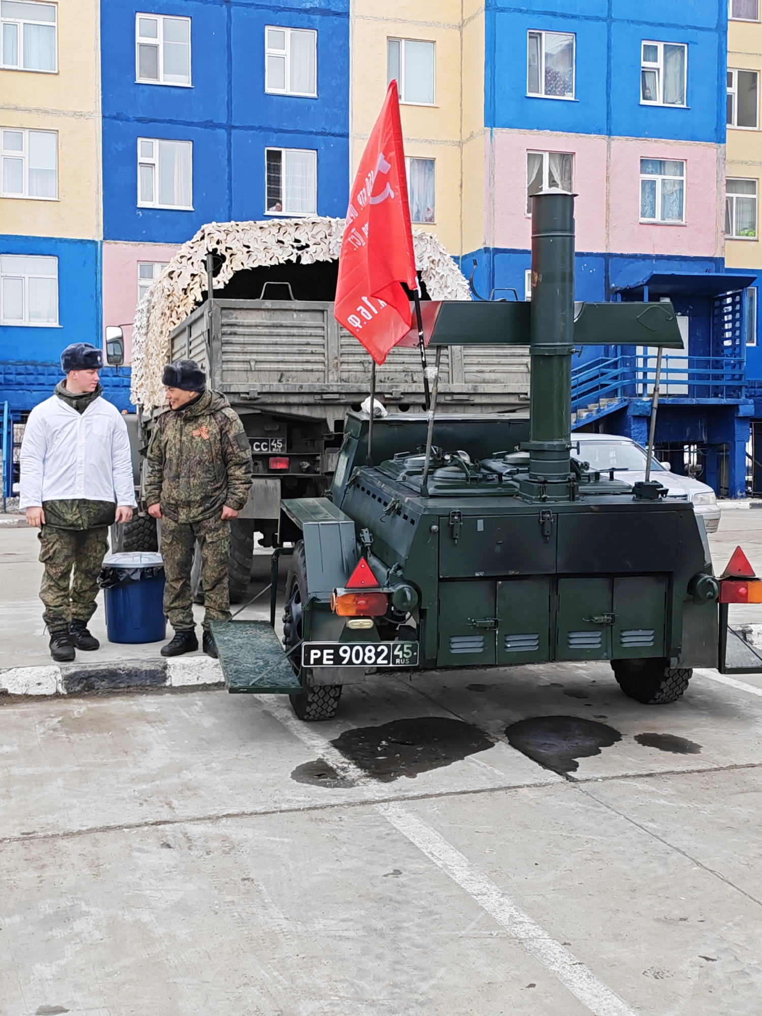 The image depicts two people in military attire standing beside a mobile field kitchen in Ugolnye Kopi, Russia. The field kitchen is mounted on a trailer, connected to a military truck camouflaged with netting. A red flag is affixed to the kitchen. The trailer's license plate reads "PE 9082 45 RUS." The buildings in the background are painted in a variety of colors: yellow, blue, and pink. One individual wears a white jacket over camouflage fatigues, while the other is fully dressed in camouflage. A blue trash bin sits beside them. A white car is partially visible behind the mobile kitchen. There are oil or water stains on the ground.