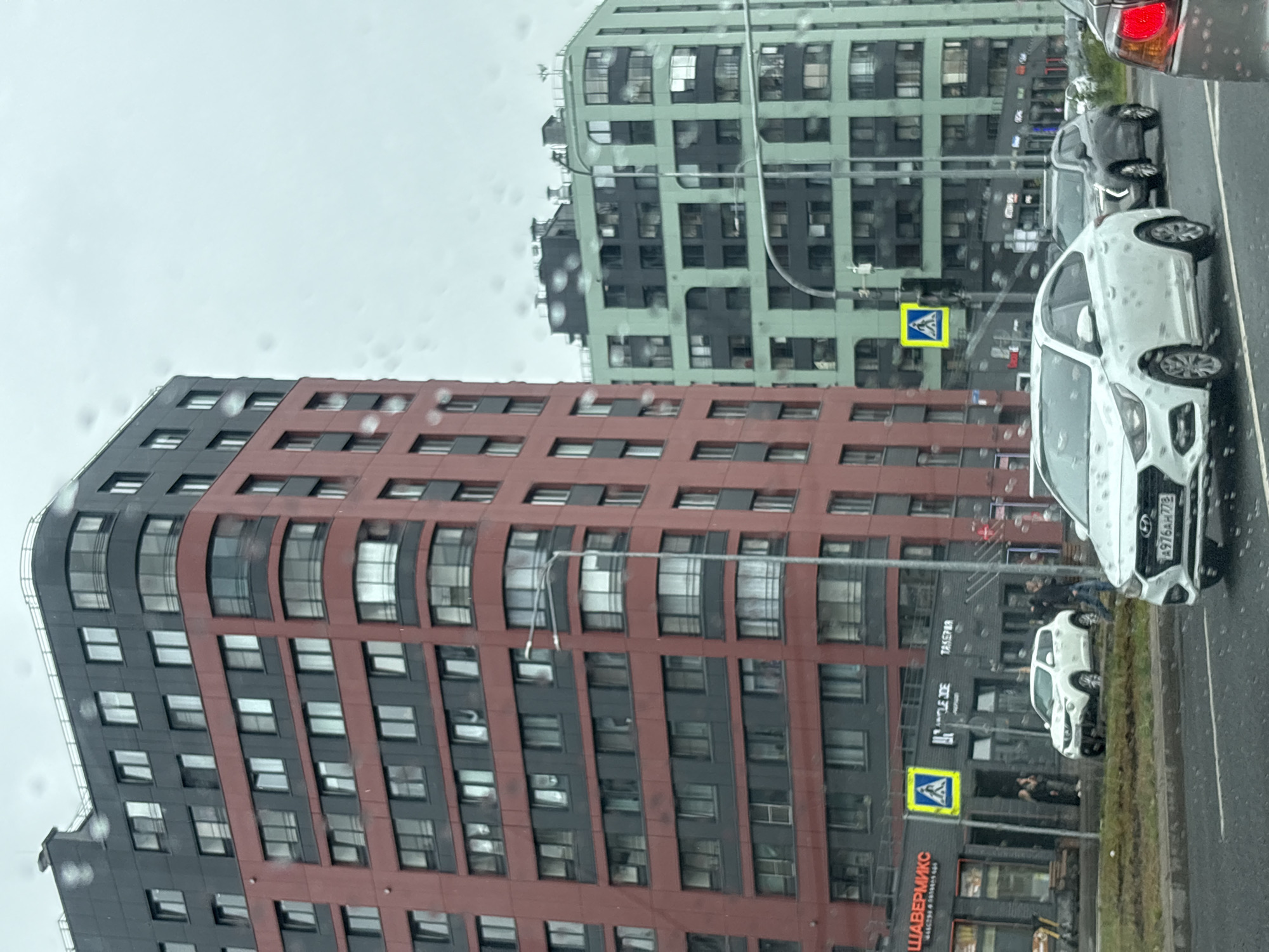 The image captures a street view in Saint Petersburg, Russia, under overcast conditions, with raindrops visible on the camera lens. Two multi-story buildings dominate the scene. The building on the left is a tall, modern residential complex with a distinctive design, featuring a red lower section and a dark grey upper section. Numerous windows are visible across its facade. To the right of this building is another contemporary apartment complex, primarily light green with dark grey accents, also featuring a multitude of windows and balconies.
In the foreground, a white sedan with the license plate A976AH 778 is in motion on a wet asphalt road. Another car, a dark-colored SUV, is visible behind the white sedan. To the left of the road, there is a sidewalk with a few pedestrians. One building at street level has storefronts with signage including "ШАВЕРМИКО" and "TAKEPAR". A yellow pedestrian crossing sign is also visible. The overall impression is of an urban environment during a rainy day.