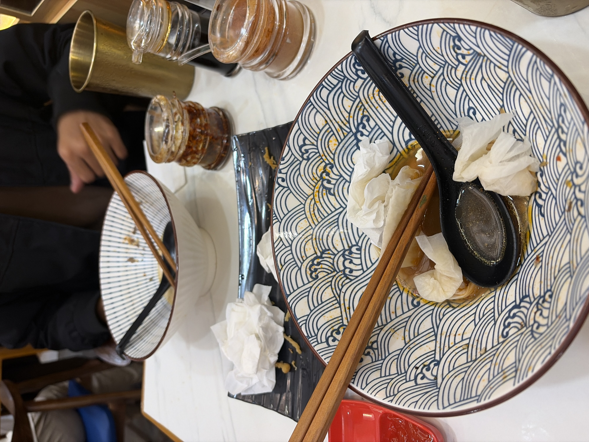 The image shows a table setting at a restaurant, likely in Pekalongan, Indonesia, with two bowls of ramen, chopsticks, spoons, and condiment bottles. The bowls are partially empty, indicating that a meal has been consumed. One bowl in the foreground has chopsticks and a black spoon resting inside, along with crumpled napkins. The other bowl in the background also contains chopsticks. Condiment bottles and a golden cup are visible on the table. A person is partially visible in the background, wearing a dark jacket. The table appears to be white with a marble pattern.