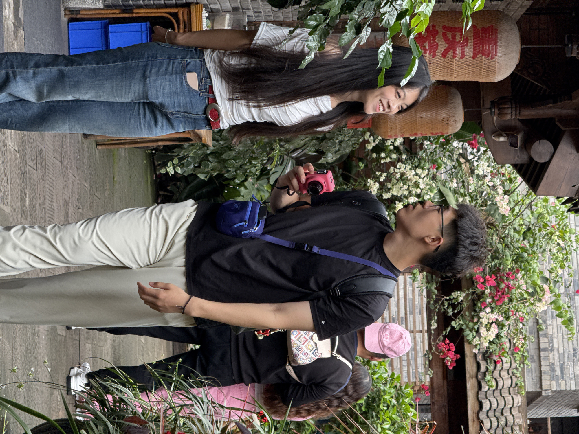 A group of four people stand outdoors in the Yuzhong District, China, taking photographs near a wall adorned with flowering plants. A young woman, positioned at the top, wears a white shirt and jeans with a red belt, her long dark hair cascading down as she looks to the side. Below her, a young man in a black t-shirt and light-colored pants holds a pink camera, aiming it towards the woman. He is wearing glasses and has a blue bag slung across his body. Further down, two more figures are partially visible; one wears a pink hat and carries a patterned white purse. Traditional Chinese lanterns hang nearby.