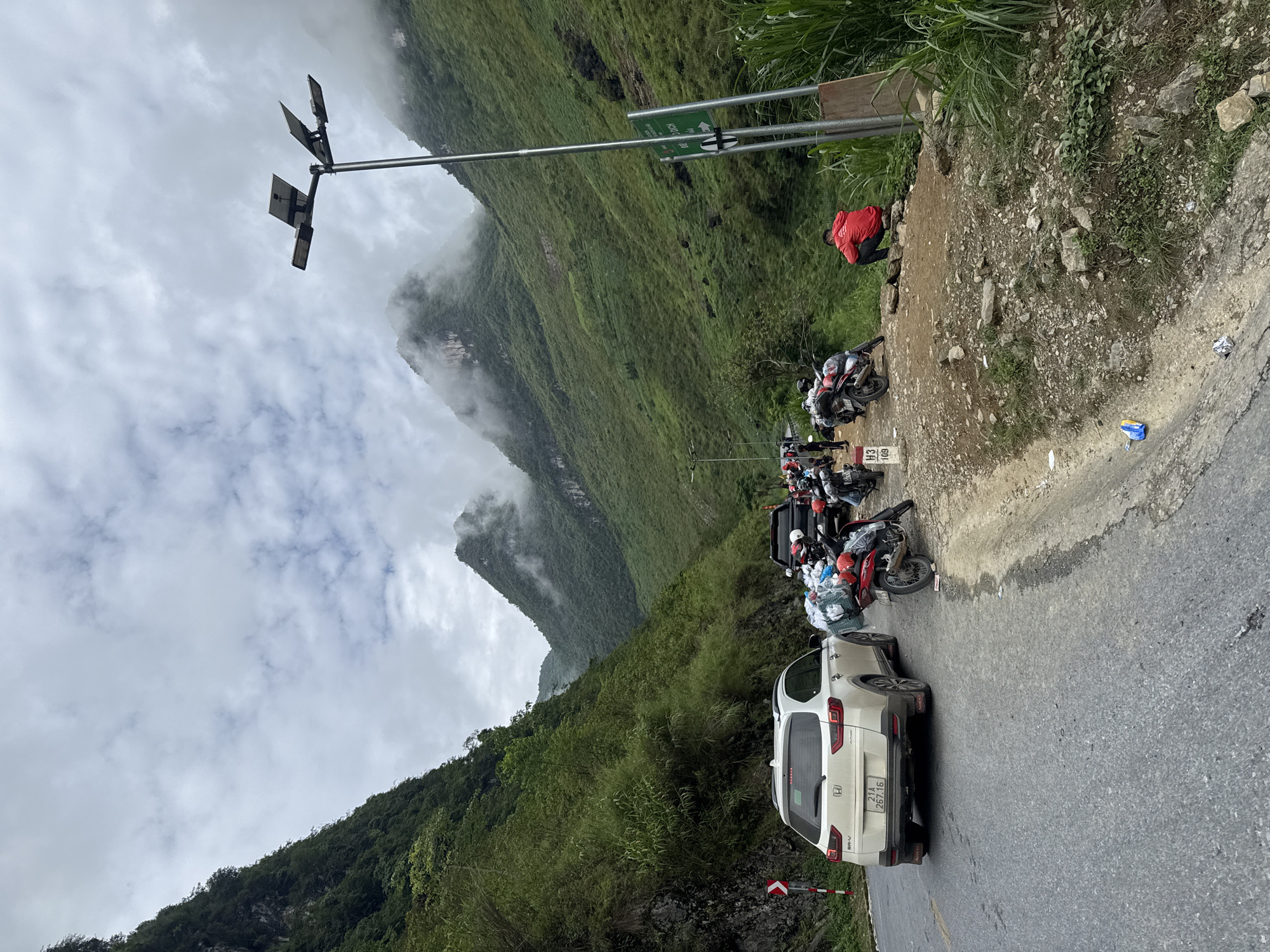 A line of parked motorcycles and a white SUV, a Honda BR-V, are on the side of a winding road in Yên Minh Commune, Vietnam. The road is next to a steep, green, mountainous terrain partially obscured by clouds. A person in a red jacket stands on the edge of the road. A tall metal pole with solar panels and streetlights is visible.