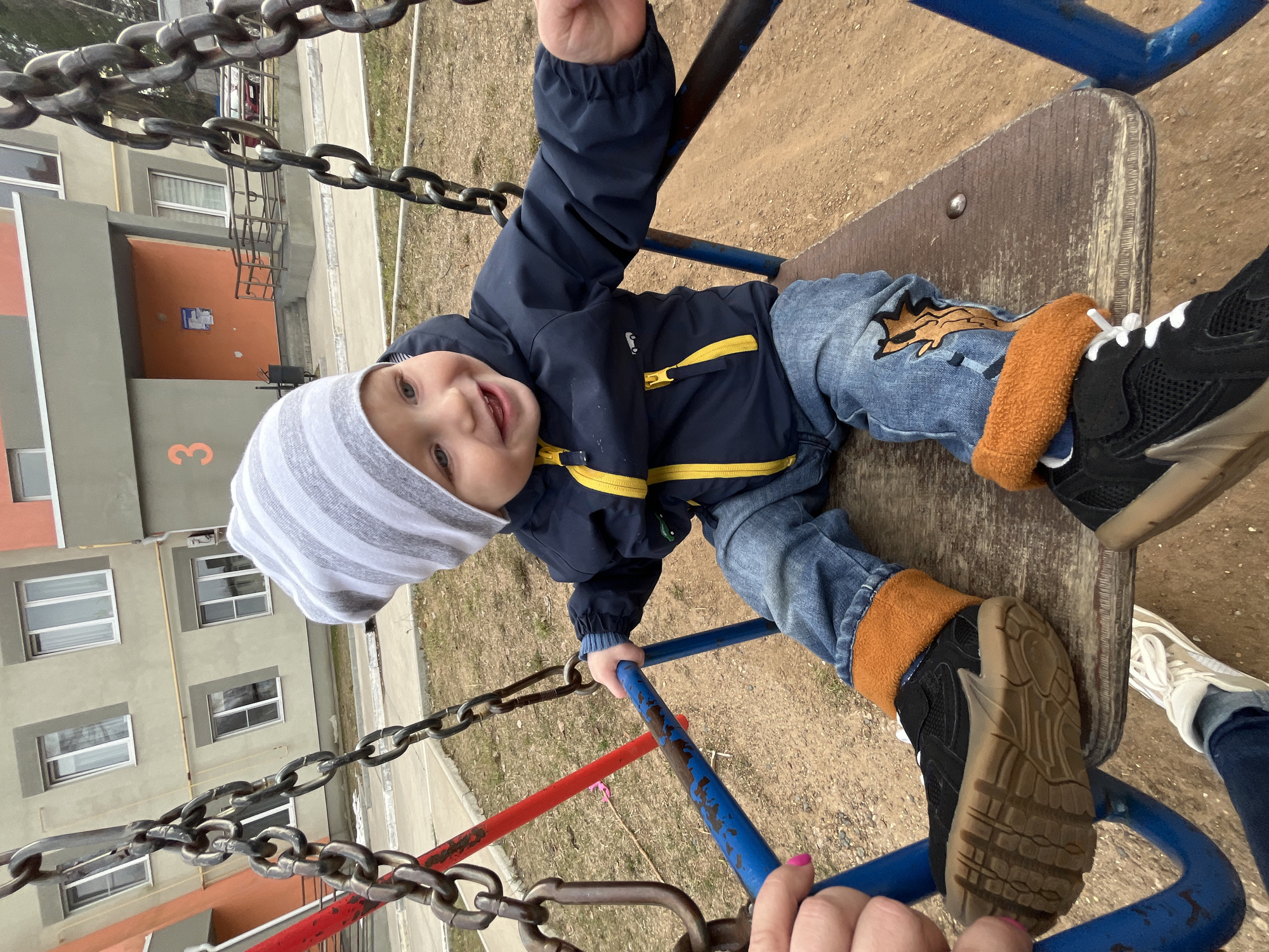 The image captures a joyful young child sitting on a swing at an outdoor playground, photographed from a low angle. The child, likely a toddler, looks directly at the camera with a wide smile. They are dressed in a navy blue jacket with bright yellow zippers, blue jeans featuring a distinct brown animal-shaped patch on the knee, a grey and white striped beanie, and black and brown sneakers with white laces. Their right hand is holding onto the blue metal frame of the swing.
At the bottom of the frame, the hands of an adult, one with visible pink nail polish, are seen holding onto the swing's structure, suggesting they are pushing the child or are also on a swing. The swing set itself features weathered blue and red metal bars, with thick metal chains suspending a worn wooden seat.
In the background, multi-story apartment buildings are visible. One building section is light grey, while another is a muted orange-brown, prominently displaying the number "3" on its facade. The ground surrounding the swing is dry, light brown dirt or sparse grass, indicating a clear, mild day, possibly in spring or early autumn. The overall scene portrays a cheerful moment of outdoor play in a residential area of Chaikovsky, Russia.