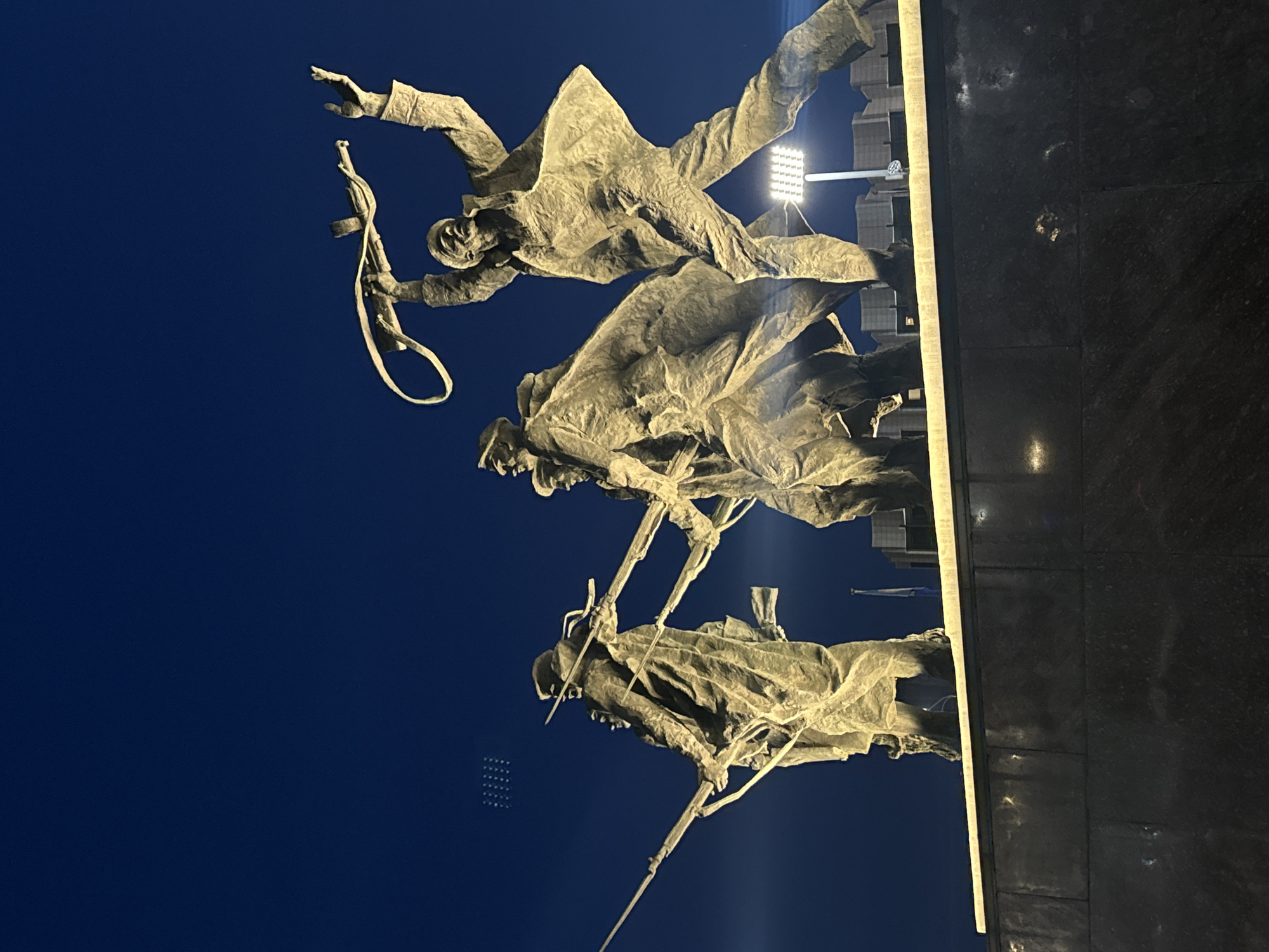 The image displays a bronze sculptural group, part of the Monument to the Heroic Defenders of Leningrad, situated at Victory Square (Ploshchad Pobedy) in Saint Petersburg, Russia. The scene is captured at night or deep twilight, with the monument illuminated by artificial lights against a deep indigo-blue sky.
Five male figures, representing soldiers or partisans, are depicted in dynamic, forward-striding poses. From left to right:
1. A partially visible figure holds a long rifle horizontally.
2. A soldier in a long coat, holding a rifle with a visible bayonet pointing upward.
3. A figure largely obscured by the second, also appearing to be a soldier in a long coat.
4. Another soldier in a long coat, moving forward, holding a rifle with a bayonet.
5. A prominent figure on the far right, wearing a jacket and trousers, is depicted in vigorous motion, raising a rifle with a sling over his head with both hands, his left leg advanced.
The statues are rendered in a dark metallic material, likely bronze, which appears a warm golden-brown due to the direct artificial illumination. They are mounted on a substantial, multi-layered plinth constructed from dark, polished stone, consistent with granite.
In the background, behind the monument, indistinct building structures are visible, along with a flagpole carrying a dark, undifferentiated flag. A bright, rectangular LED floodlight mounted on a pole provides illumination from the lower right of the sculptural group. Faint, grid-like patterns of light are discernible in the upper left portion of the dark sky, suggesting reflections or distant light sources.