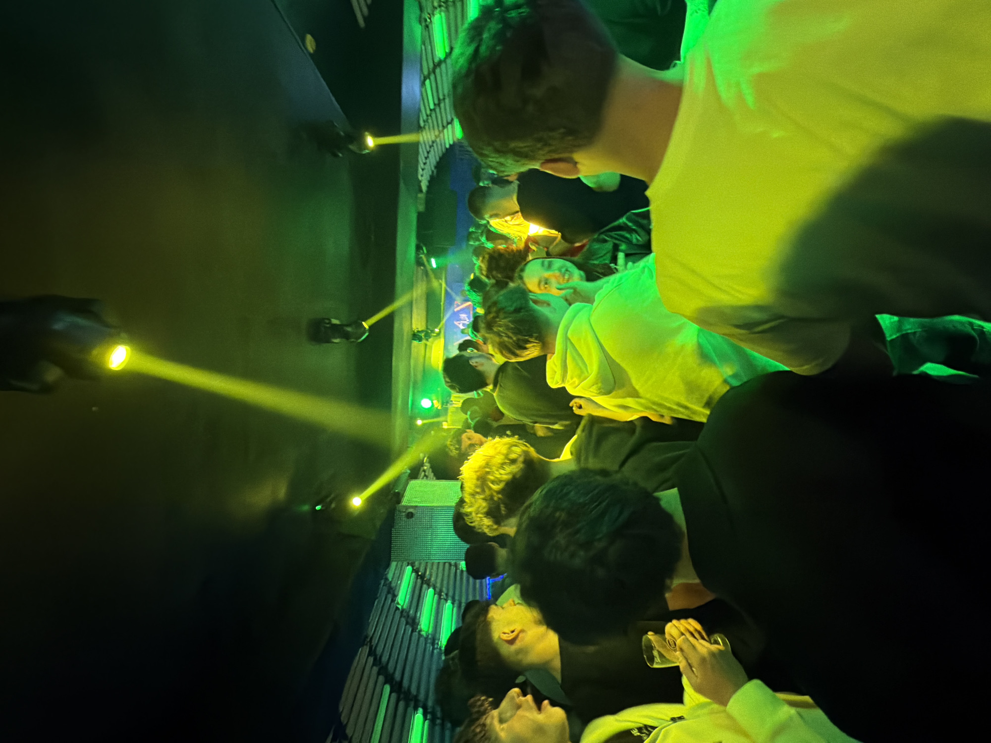 The image shows a crowded indoor event, possibly a concert or club, likely in Vila Real, Portugal. People are standing close together, illuminated by green and yellow stage lighting. Some individuals are looking up, possibly toward a stage or performance area. At least one person is holding a glass. The environment is dark with visible stage lights mounted on a wall or ceiling. There are a few people on stage.