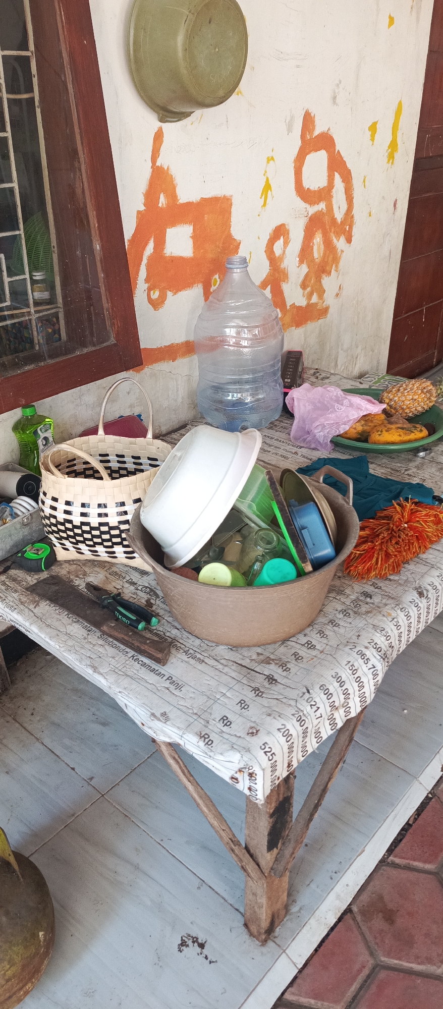 An outdoor scene is depicted with various objects placed on a table covered with a sheet of paper featuring monetary denominations. In the foreground, on the left, a large, green, round object, possibly a coconut, rests on the floor. Above it, a rough wooden table has a metal container holding a measuring tape, a pair of pliers on a wooden block, and a woven basket with a checkered pattern. To the right of the basket is a large, brown plastic basin filled with dishes, including a white bowl and several green and clear plastic containers. An orange duster is positioned to the right of the basin. In the background, a large, clear plastic water jug sits on the table. To its left, a green bottle of liquid is visible. The wall behind the table is painted white with orange and yellow abstract designs. A window with a green screen is to the left of the water jug. The table is situated on a tiled floor. The context of Arjasa, Indonesia, suggests this is likely a residential or workshop area.