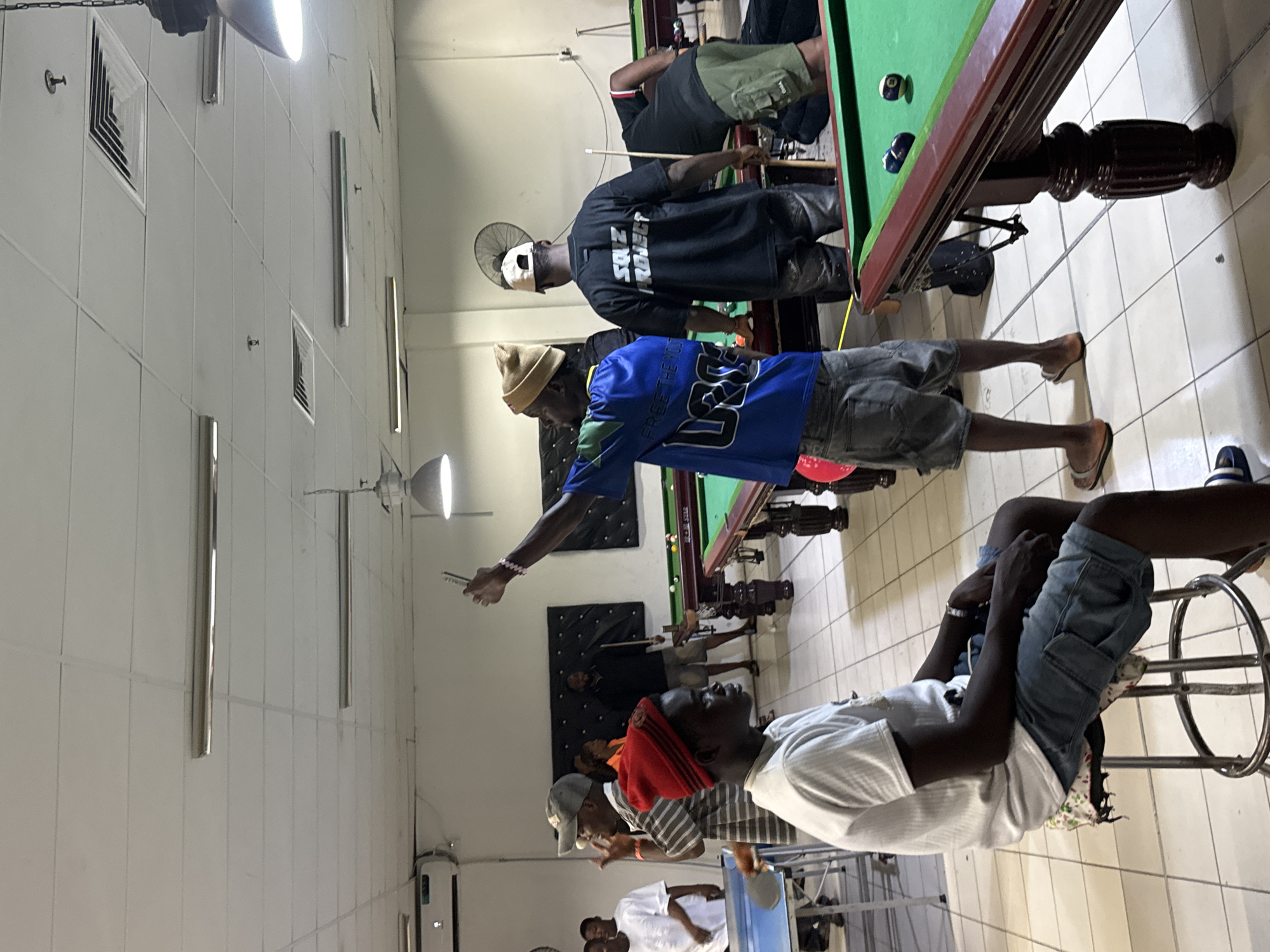 **Location:** The image depicts an indoor pool hall in Onitsha, Nigeria.
**People:** Several African men are present. Some are actively playing pool, while others are observing or sitting. One man is wearing a blue shirt with "FREE THE 107" printed on it. Another man is wearing a black shirt with "SQZ PROJECT" printed on it.
**Objects:** A pool table dominates the scene. Pool cues, balls, and chairs are also visible.
**Actions:** Men are playing pool. Other actions include watching the game and sitting.
**Interactions:** Players are interacting with the pool table and equipment. There are interactions between the people watching and playing.
**Environment:** The pool hall has tiled flooring and white walls with visible air conditioning units. Fluorescent lighting illuminates the space.