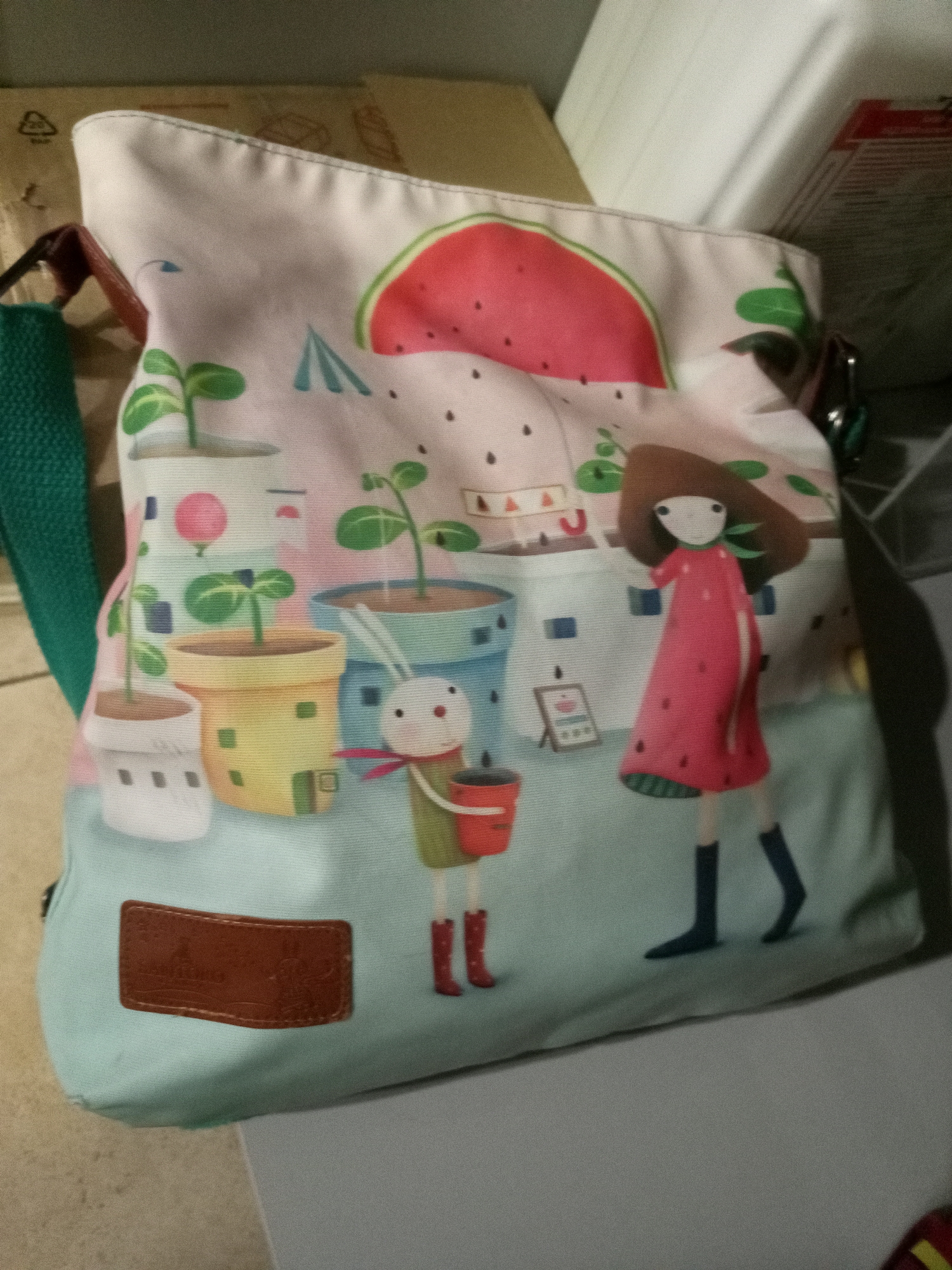 This is a close-up, eye-level, indoor photograph of a canvas tote bag with a light blue bottom and a white upper portion. The bag features a whimsical print of a cartoon garden scene. The print includes a large slice of watermelon at the top, stylized buildings with windows, potted plants with green sprouts, a rabbit character in red boots holding a red bucket, and a young girl with dark brown hair wearing a pink dress and dark blue boots. A brown leather tag with embossed text is affixed to the bottom front of the bag. The bag is placed on a light gray surface, with other objects in the background, including what appears to be cardboard boxes. The lighting suggests the photo was taken indoors with artificial light.