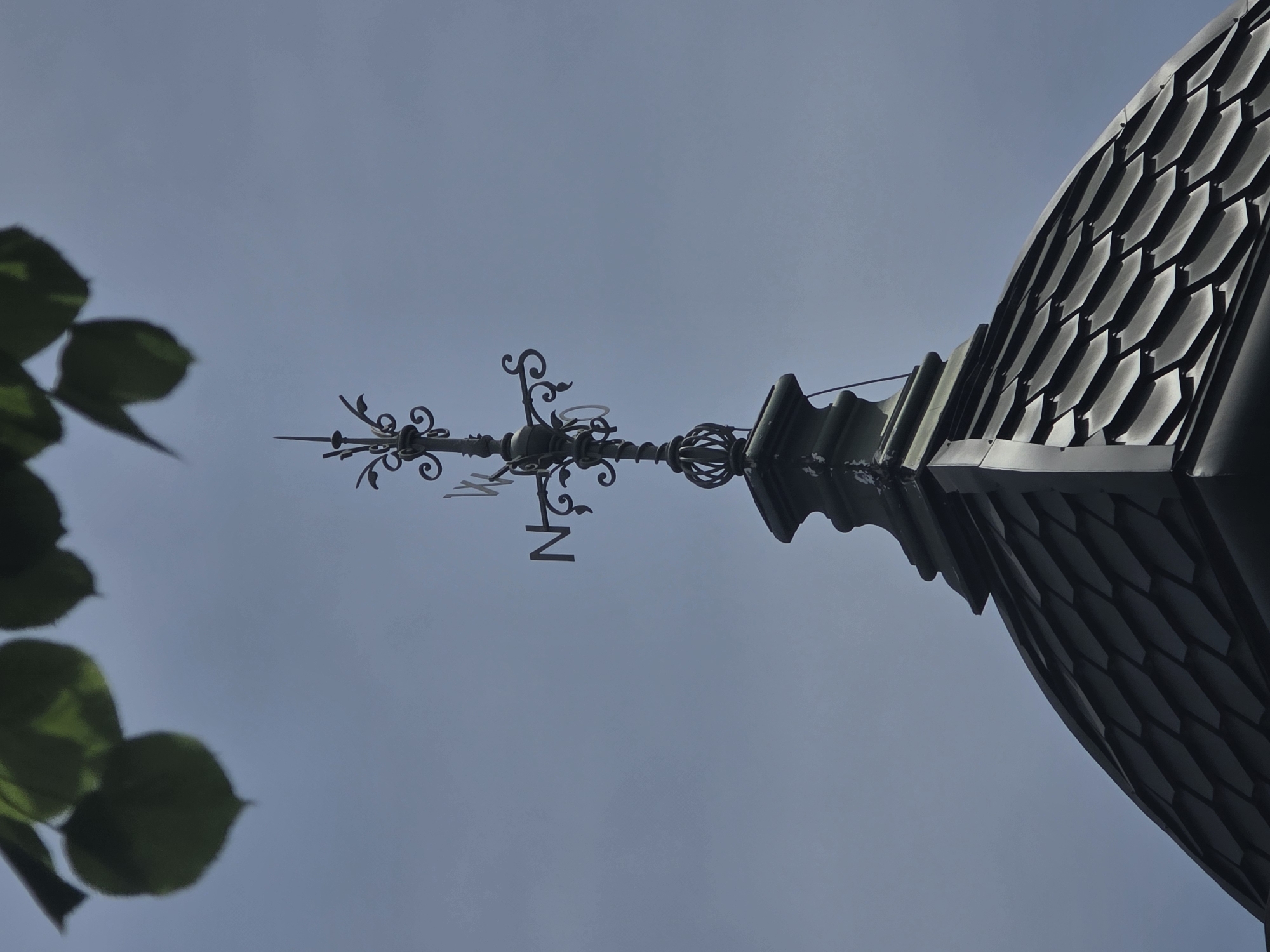 A low-angle shot reveals a detailed weather vane atop a building, likely in Krasnodar, Russia. The weather vane, composed of ornate metalwork, features a directional "N" for North. The roof, covered in dark, scale-like tiles, curves upward to meet the vane. Soft green leaves are visible on the left edge of the frame, adding a touch of foliage to the urban scene. The sky is a pale blue, devoid of clouds, emphasizing the architectural details.