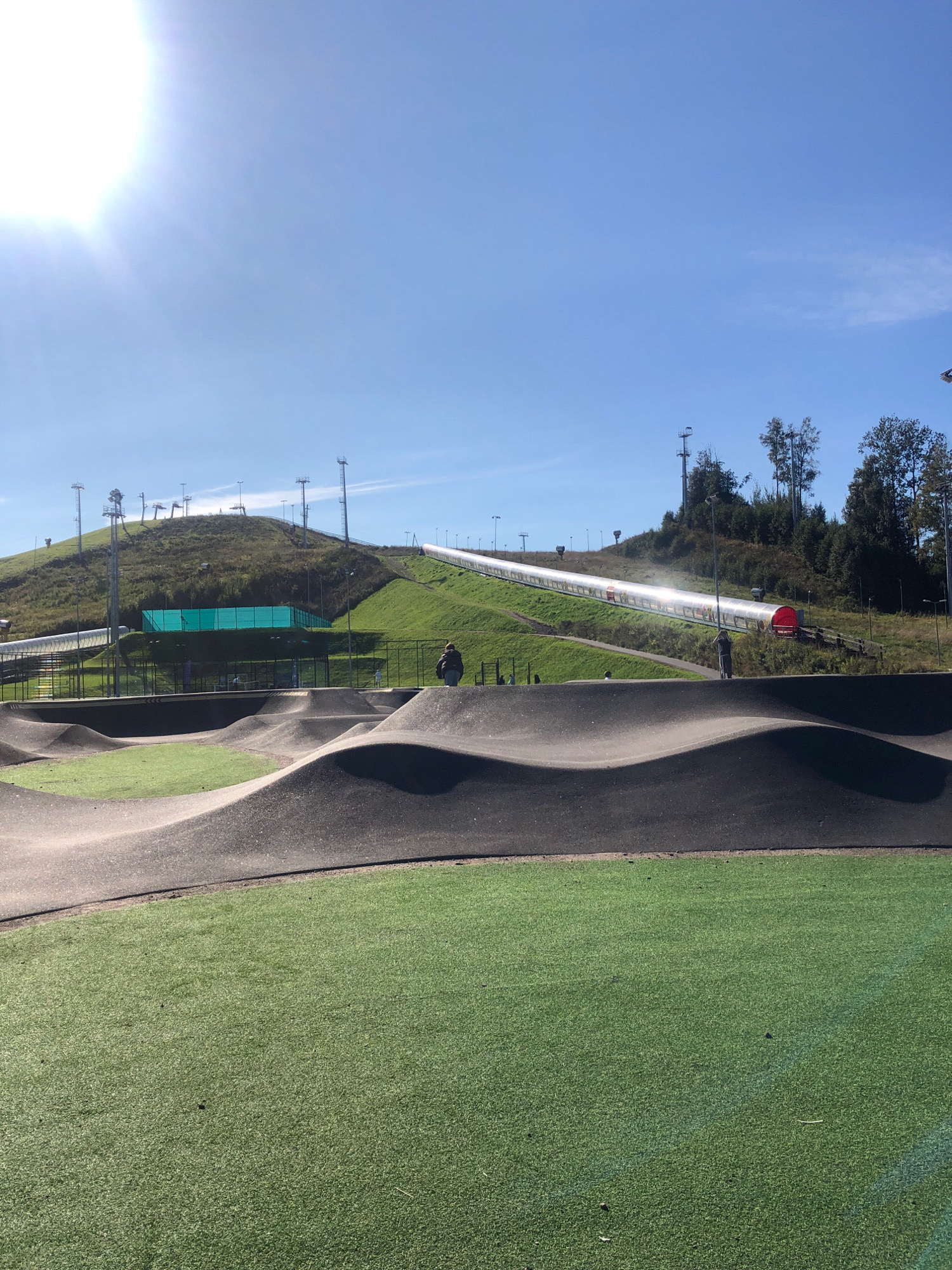 This is a photograph of a recreational area in Okhtinskiy Park, Russia, on a sunny day. In the foreground, a pump track with undulating, dark gray surfaces is visible, bordered by artificial turf. Beyond the pump track, a green artificial turf field is present. In the mid-ground, a long, enclosed tube slide with a transparent upper section runs down a grassy hill. There are people visible both on the pump track and near the entrance to the tube slide. The background consists of a green, grassy hill dotted with trees and several tall light poles and ski lift towers. The sky is bright blue with a prominent sun in the upper left corner.