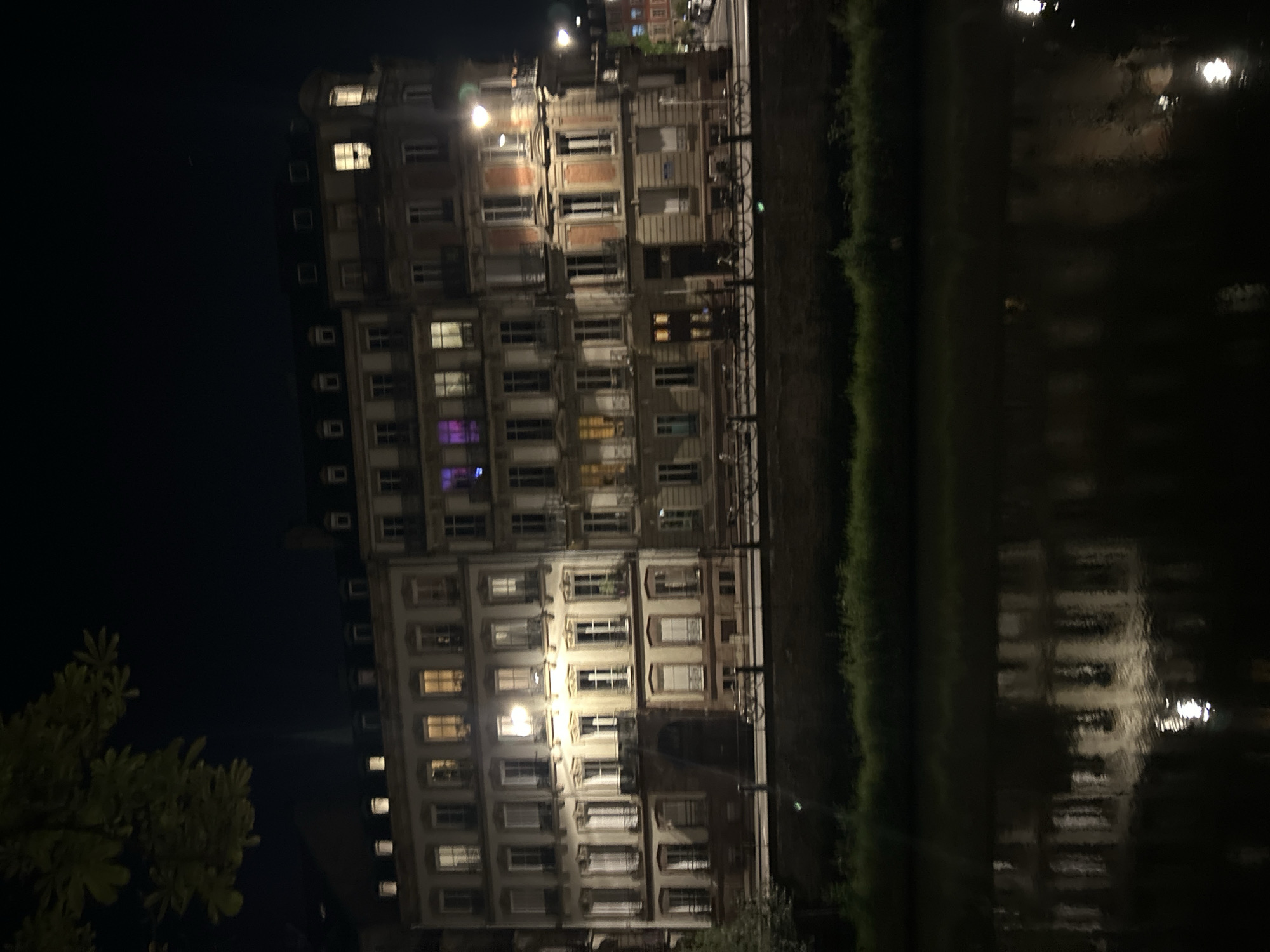 The image depicts a multi-story building in Strasbourg, France, at night, reflected in a body of water. The building has numerous windows, many illuminated. Vegetation lines the edge of the water. The overall lighting is dim.