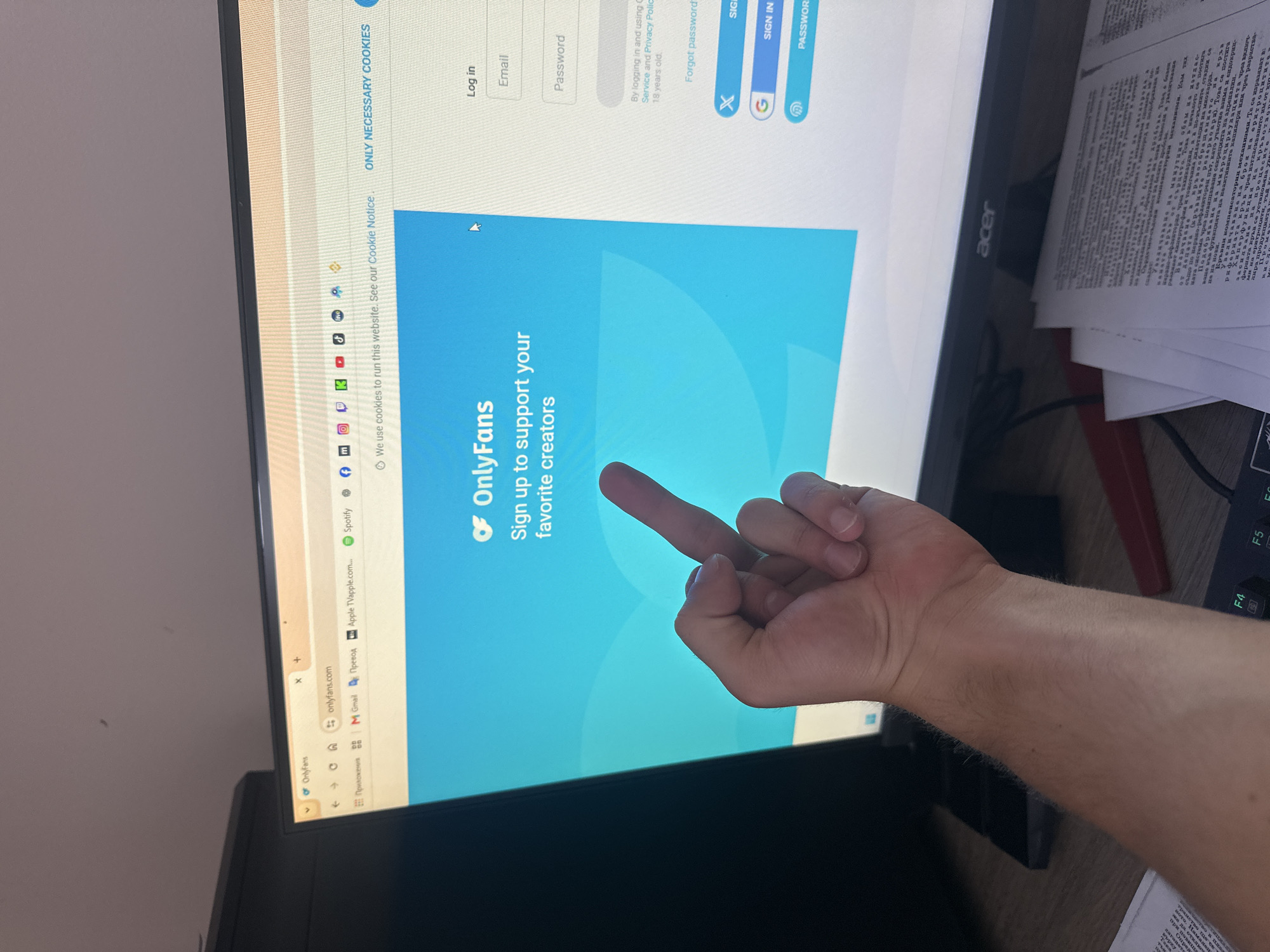An indoor personal workspace, identified as being located in Saraia, Bulgaria, is depicted from a first-person perspective. In the foreground, a light-skinned forearm and hand are prominently positioned, extending from the lower right towards the center of the frame. The hand is engaged in an action, displaying a middle finger gesture directly towards a computer monitor.
The central subject is a black Acer-branded computer monitor, displaying a web browser interface open to "onlyfans.com". The webpage features a blue background with the white "OnlyFans" logo and text stating, "Sign up to support your favorite creators." To the right, a login panel includes fields for "Email" and "Password," alongside "Log in" options via X (formerly Twitter), G (Google), and a password manager icon. A cookie notice is visible across the top of the main content area, stating, "We use cookies to run this website. See our Cookie Notice." The browser's top bar displays a tab for "OnlyFans" and various bookmarks, including "Приложений" (Applications), "M Gmail," "Перевод" (Translate), "Apple TVapple.com," "Spotify," and icons representing social media platforms such as Facebook, Instagram, Twitch, TikTok, YouTube, and Binance.
The monitor rests on a dark wooden desk. To the right of the monitor and in the foreground, multiple sheets of white paper with black text, appearing to be in Cyrillic script, are stacked. A black keyboard with some green keycaps (F4, F5, F6 visible) is partially visible at the bottom left. A dark, rectangular electronic device, possibly a smartphone or external drive, lies on the desk to the left of the visible hand. The background reveals a plain, light-colored wall above the monitor. The overall lighting is even, suggesting an adequately lit room.