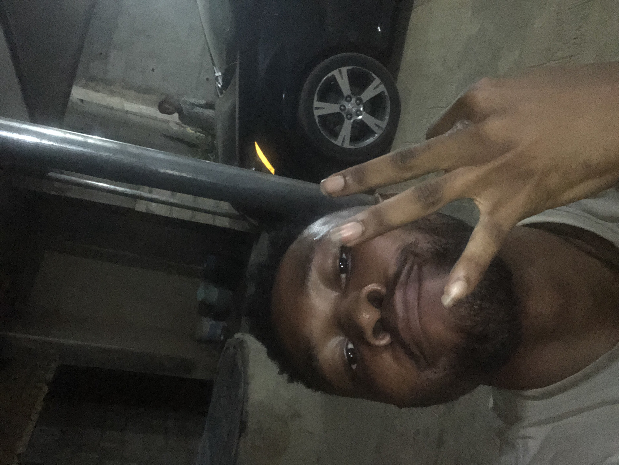 A man with dark skin and short, dark hair poses in a dimly lit environment, making a peace sign with his right hand. He has a beard and is wearing a white t-shirt. Behind him, there's a black car with a silver rim wheel. A large metal pipe or beam stretches across the left side of the frame. The setting appears to be an underground or dimly lit parking garage. The location is Unknown, Unknown.