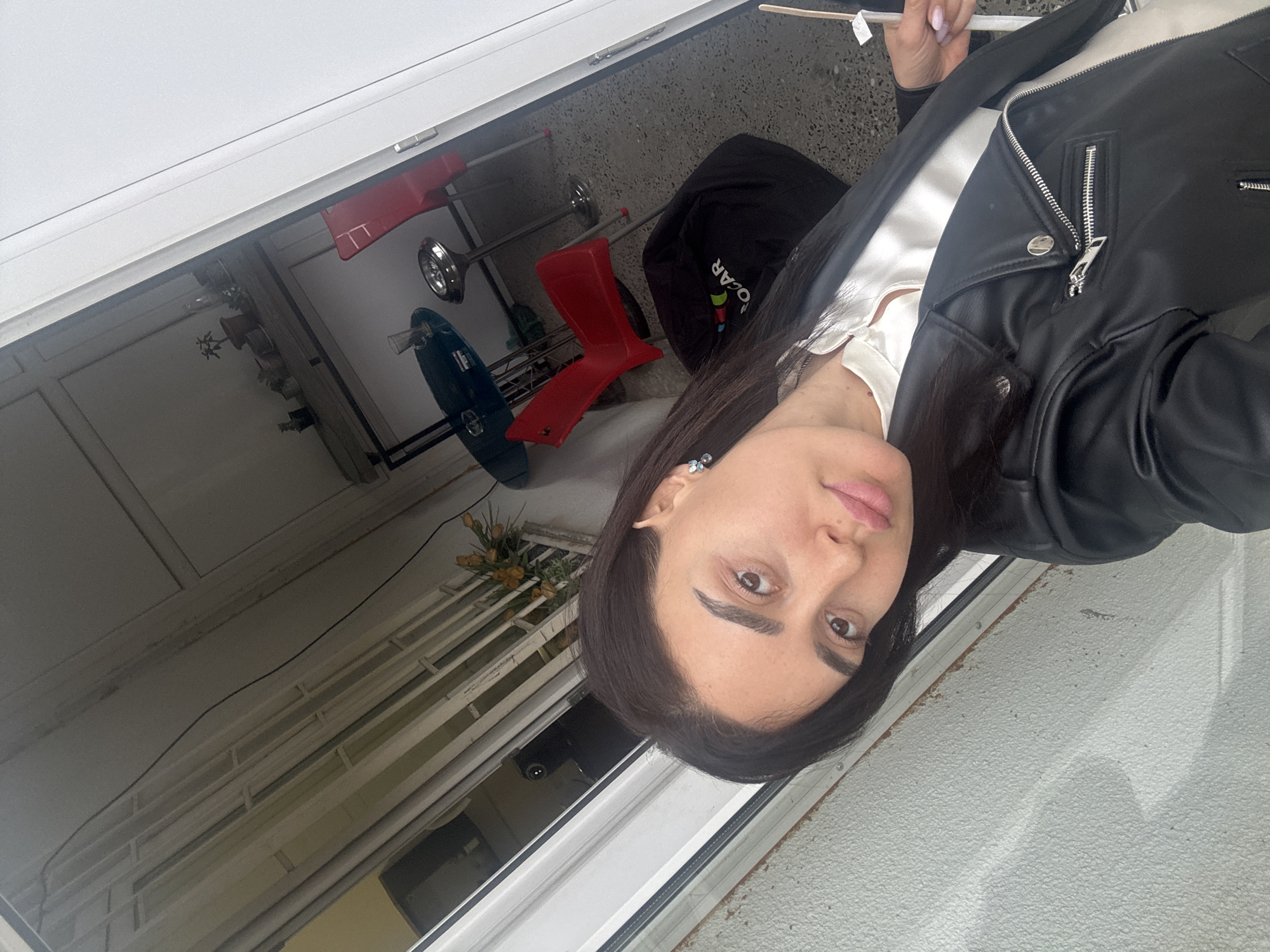 A woman in a black leather jacket is taking a selfie. She is wearing a white top. In the background, there is a red and blue child's toy, a black bag with a colorful logo, and a pot of yellow flowers. The location appears to be an outdoor patio or balcony, possibly in Tbilisi, Georgia, based on architectural styles common to the region. The lighting indicates it's daytime.