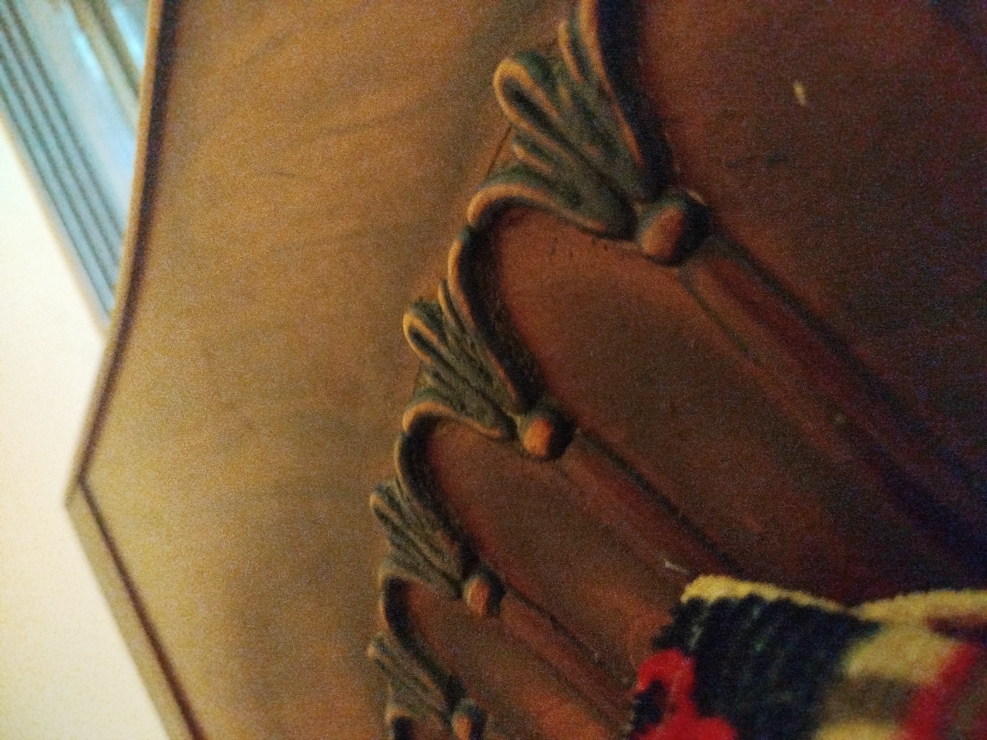 The image is a close-up, low-angle shot of decorative wooden carvings on a piece of furniture. The carvings are ornate, with a fan-like or leaf-like design and appear to be painted a dark green with lighter, rounded accents in a peachy-orange color. The furniture itself is made of a dark wood with a reddish-brown hue. In the lower right corner, a portion of a colorful textile with red, yellow, and dark blue patterns is visible, partially overlapping the wooden carving. The background is a textured, light brown surface, possibly wood grain, with a blurred bright area on the left edge that could be light filtering through a window or reflecting off a surface. The image is grainy and out of focus in some areas, suggesting it was taken with a low-resolution camera or in poor lighting conditions. The scene is indoors, with no clear indication of an outdoor environment or specific location.