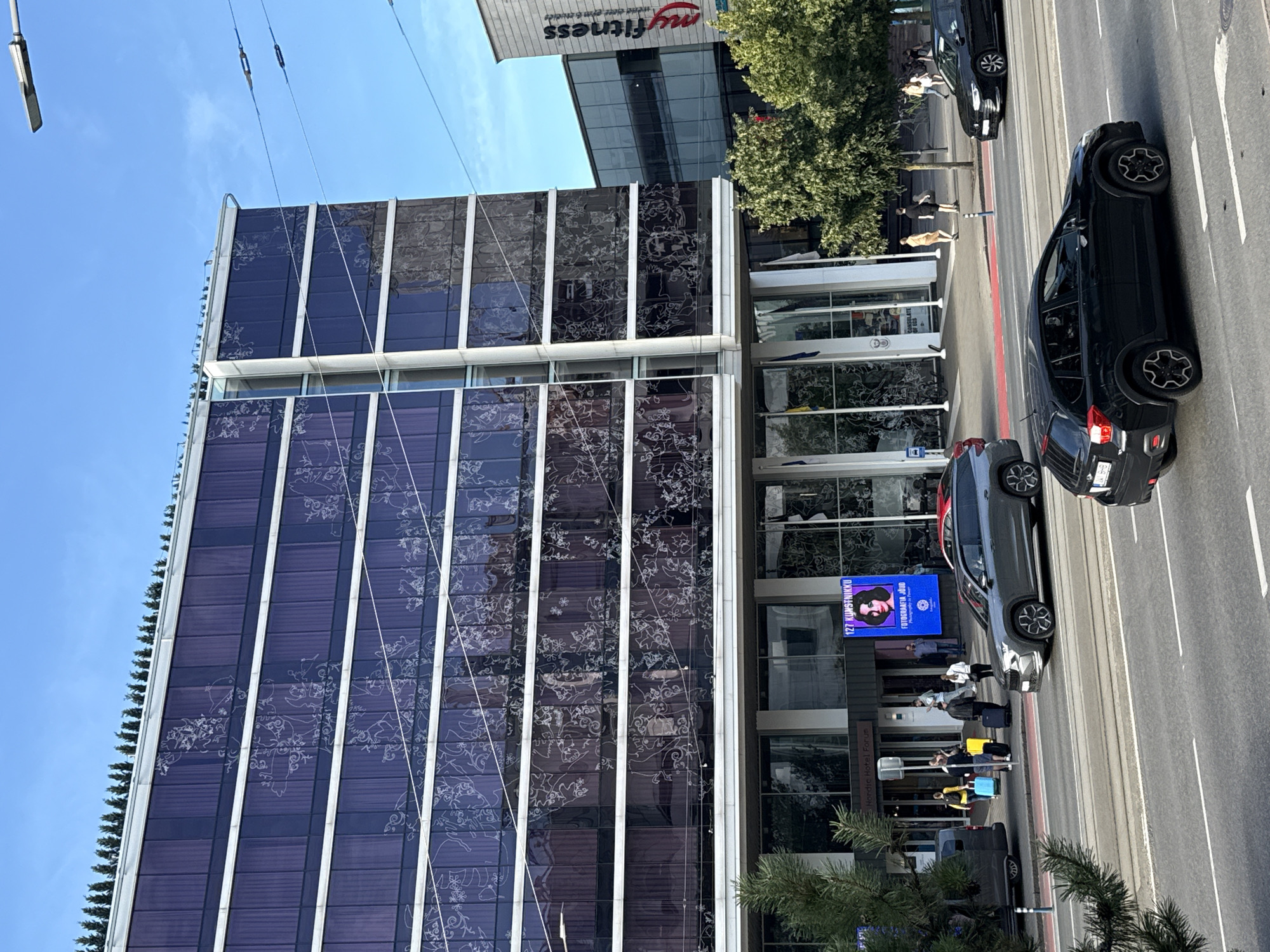 A wide, eye-level, outdoor, daytime shot captures a street scene in front of a modern, multi-story building with dark purple, reflective glass facades adorned with white, intricate, floral-like patterns. The building has a flat roof topped with a dense row of evergreen trees. Several horizontal white bands divide the glass panes. Below the glass facade, a glass-enclosed ground floor features an entrance to "Nordic Hotel Forum." To the right of the hotel, a building with a white facade displays the red "myfitness" logo.
On the street in the foreground, several vehicles are present. A dark gray SUV is parked in the right lane, while a black SUV is in the center lane, moving away from the camera. Another black car is visible in the right lane, further down the street. A red car is partially visible behind the gray SUV.
On the sidewalk in front of the hotel, pedestrians are seen with luggage. One person is pulling a yellow suitcase, another is pushing a black suitcase, and a third person is carrying a blue suitcase. Other individuals are walking in various directions. A large digital screen displays a woman's portrait and text. Trees line the sidewalk. The street features multiple lanes with tram tracks. The sky is clear and blue, with visible power lines crossing the upper portion of the image.