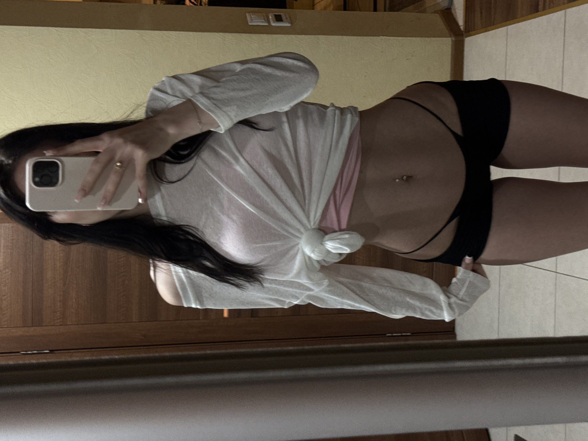 A person is standing in front of a mirror, taking a selfie. They are wearing a white off-the-shoulder top tied at the waist, a pink bralette, and black shorts. The background appears to be an indoor setting, possibly a home, with a wooden door and a yellow wall. There are tiles on the floor and a light switch on the wall. The person has long, dark hair and a navel piercing. The image quality is moderate, with some graininess and low light. The exact location within Khimki, Russia, cannot be determined from the visual information provided.