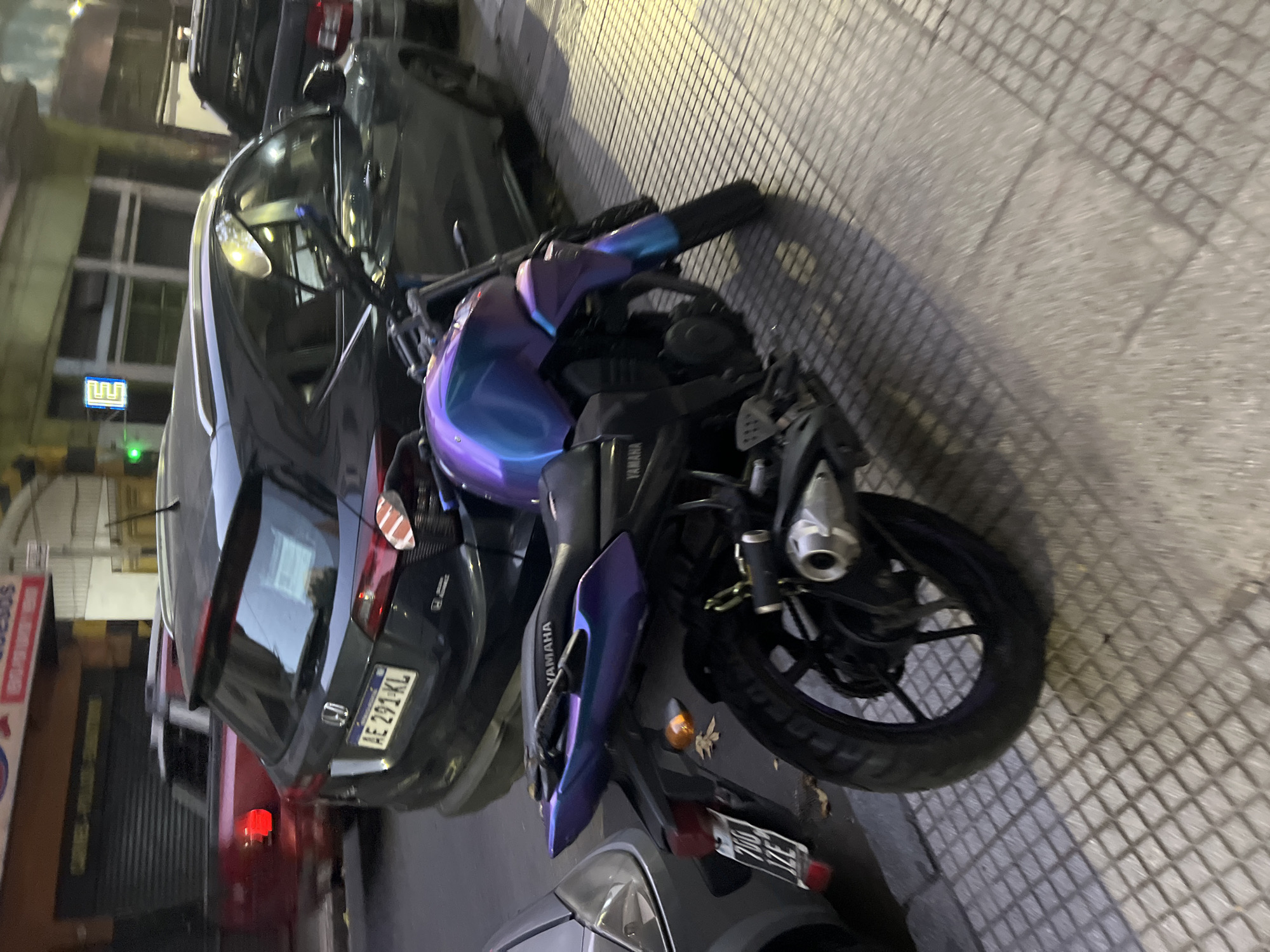 The image displays an urban street scene in Buenos Aires, Argentina, captured during the evening or night as evidenced by the artificial lighting and dark ambient conditions. The primary subject is a parked motorcycle in the foreground, which exhibits a distinctive iridescent purple and blue finish on its fairing. The brand "YAMAHA" is visible on the motorcycle's seat or side panel.
The motorcycle is positioned on a sidewalk characterized by a textured grid pattern. Behind it, a dark grey Honda CR-V is parked, showcasing an Argentine license plate that reads "AE 291 KL." A red vehicle is partially visible further in the background.
The setting includes buildings lining the street. One building features a glowing green sign that resembles a stylized "W" or "M." Another building displays a red sign with partially visible text that appears to include the word "CURESOS." No people are visible in the frame, and all vehicles appear stationary. The artificial lights illuminate the scene, casting subtle shadows across the sidewalk and vehicles.