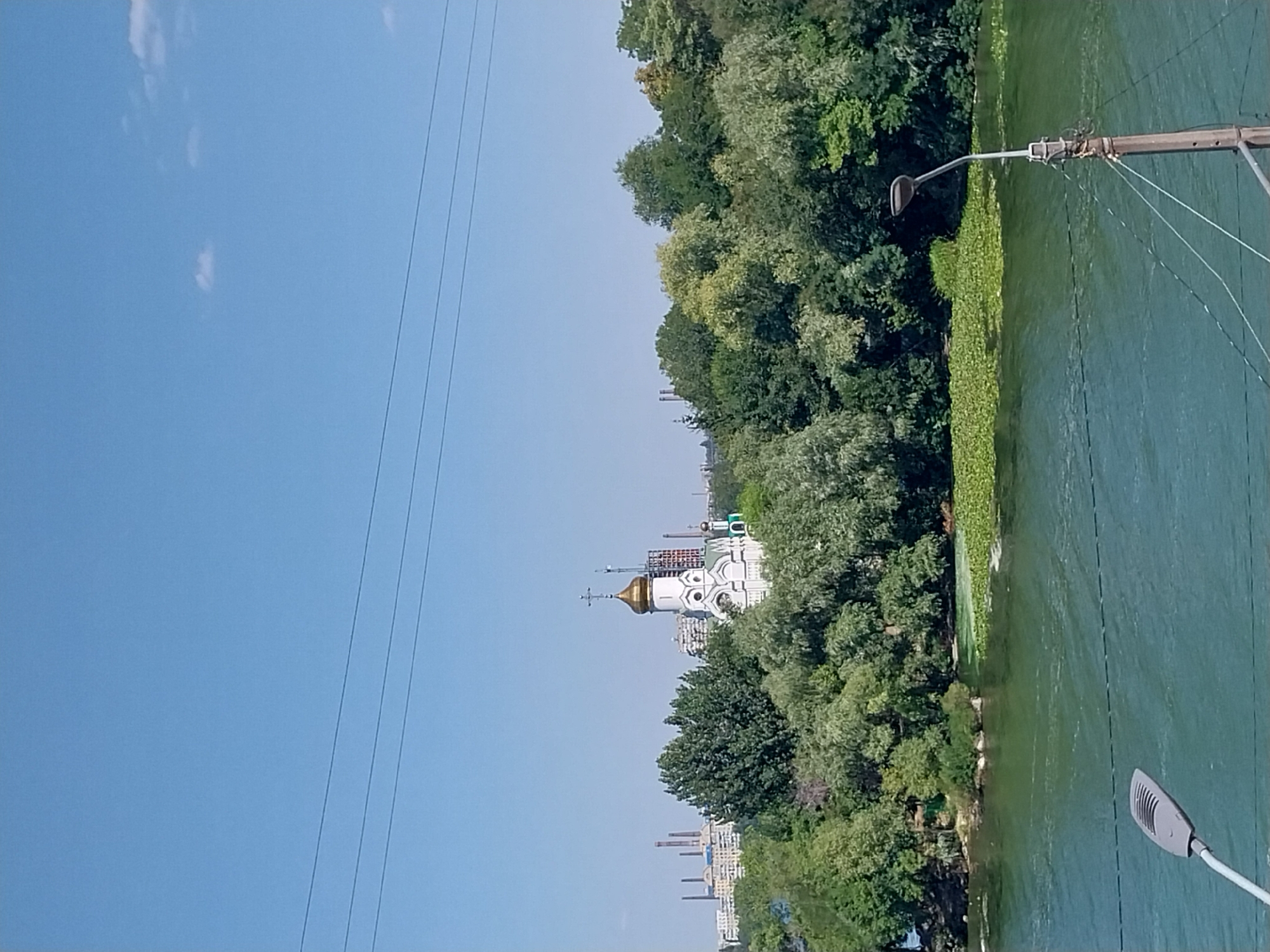 The image depicts a view across a body of water towards a church and industrial buildings in the distance. In the foreground, a portion of a modern street lamp is visible on the right, with its head pointing towards the viewer's left. The water in the mid-ground is a vibrant green and covered with lily pads in patches. A dense line of lush green trees borders the water.
Beyond the trees, a white church with a golden dome and a cross on top is prominently situated. Behind the church, several tall, smokestack-like industrial structures are visible, suggesting an industrial area of the city. The sky is a clear, light blue with a few wispy clouds. Overhead, three dark, horizontal power lines are stretched across the frame.
This scene is likely a view of the Dnipro River, potentially near the Dnipro Industrial Zone, given the presence of both a church and industrial facilities. The foliage suggests a warmer season, likely summer or late spring, with the ample greenery and clear blue sky. There are no people visible in the image. The overall impression is a juxtaposition of natural beauty, religious architecture, and industrial infrastructure.