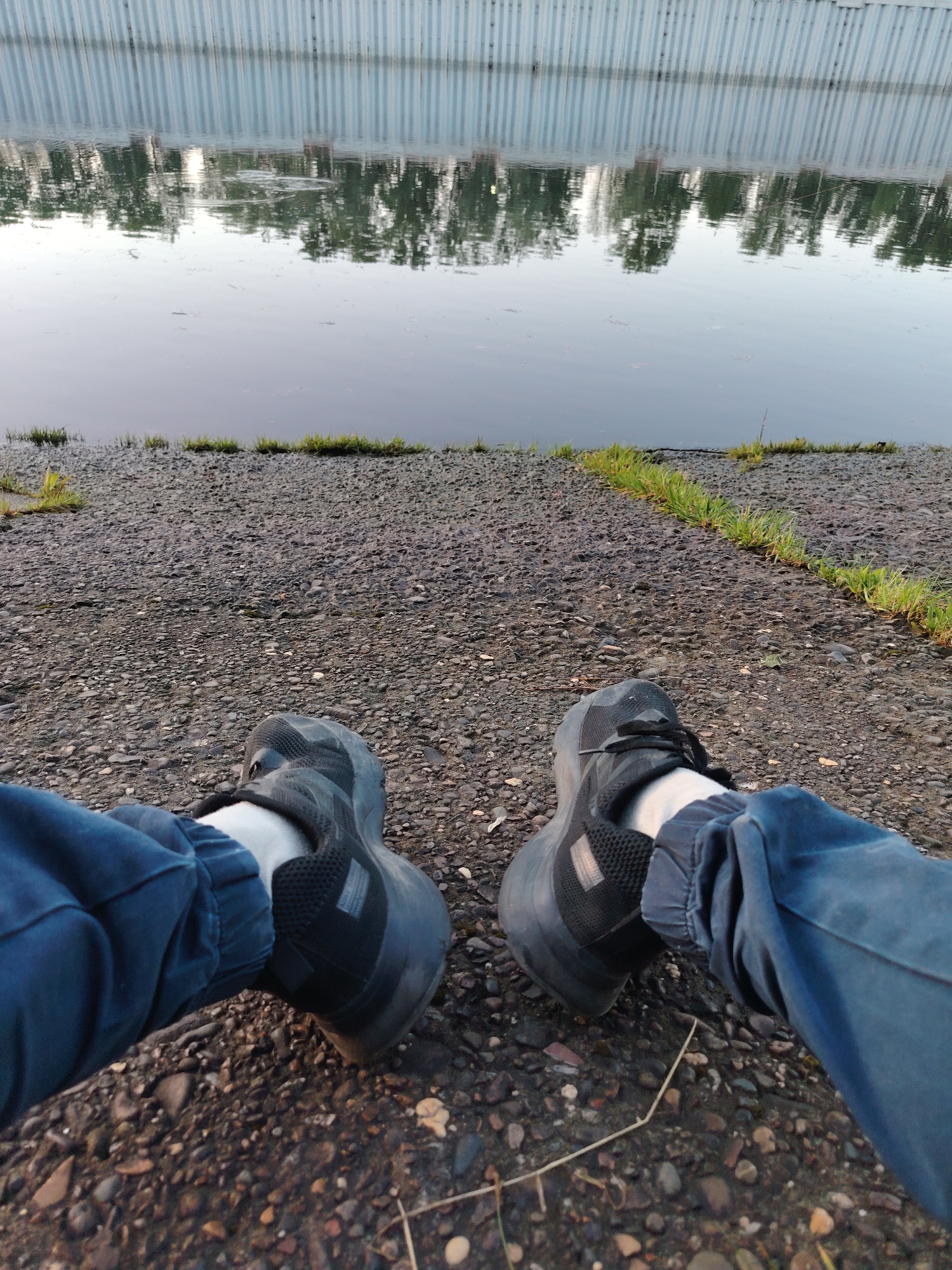 A low-angle shot shows a person seated on a pebbled ground near a body of water, likely a river or canal, in Chaikovsky, Russia. The person's legs, clad in blue pants and black athletic shoes, are positioned towards the camera. The waterway is behind the person and a concrete wall reflecting trees in its still waters, suggesting an urban setting with natural elements. The ground has patches of green vegetation.