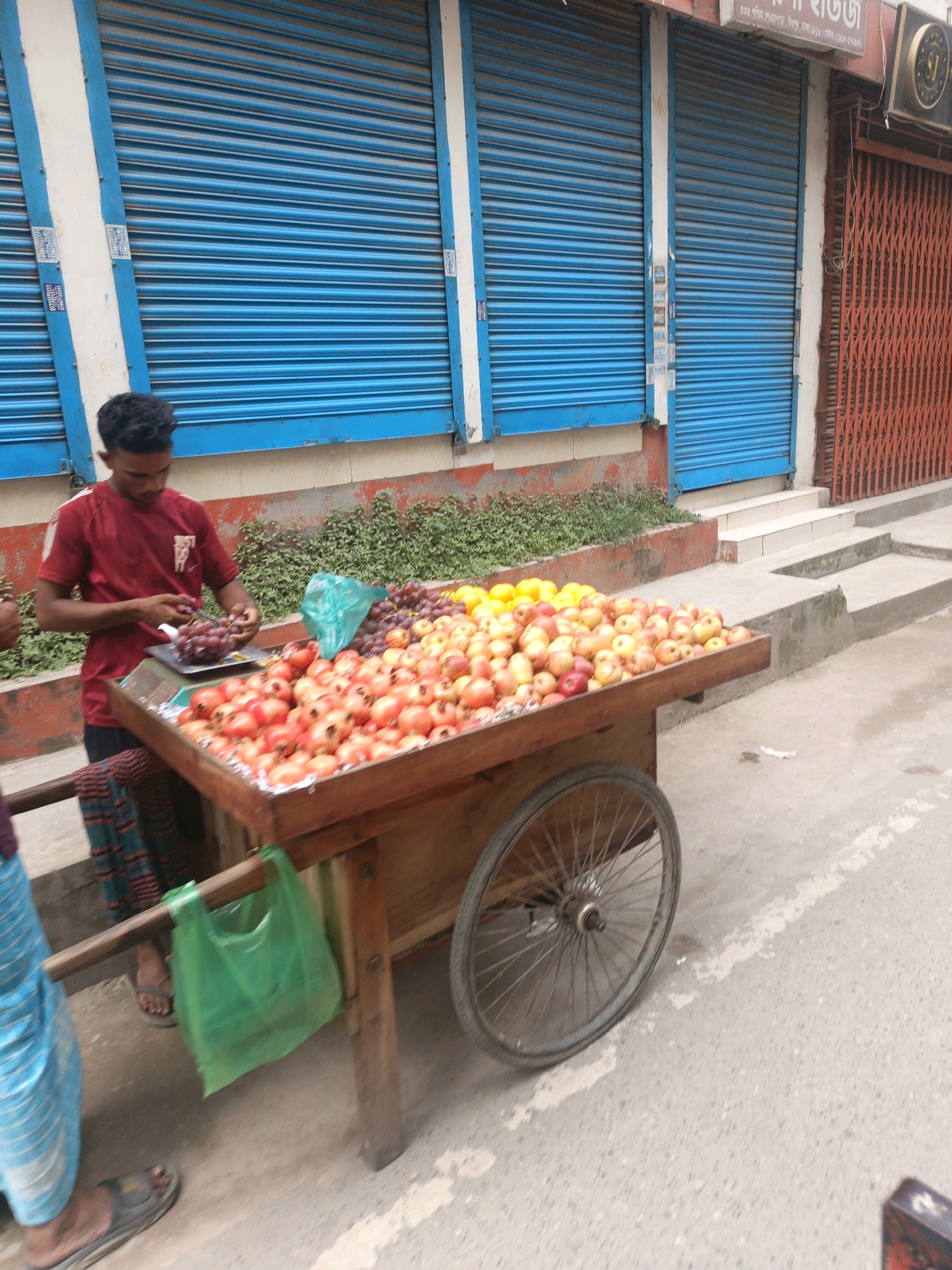 A young Bangladeshi man in a red t-shirt is preparing grapes at a fruit cart in Dhaka, Bangladesh. The cart is laden with pomegranates and other fruits. In the background, blue metal roller shutters are visible, suggesting a storefront. The man's clothing and the general setting are consistent with a street-level vendor in a bustling urban environment.