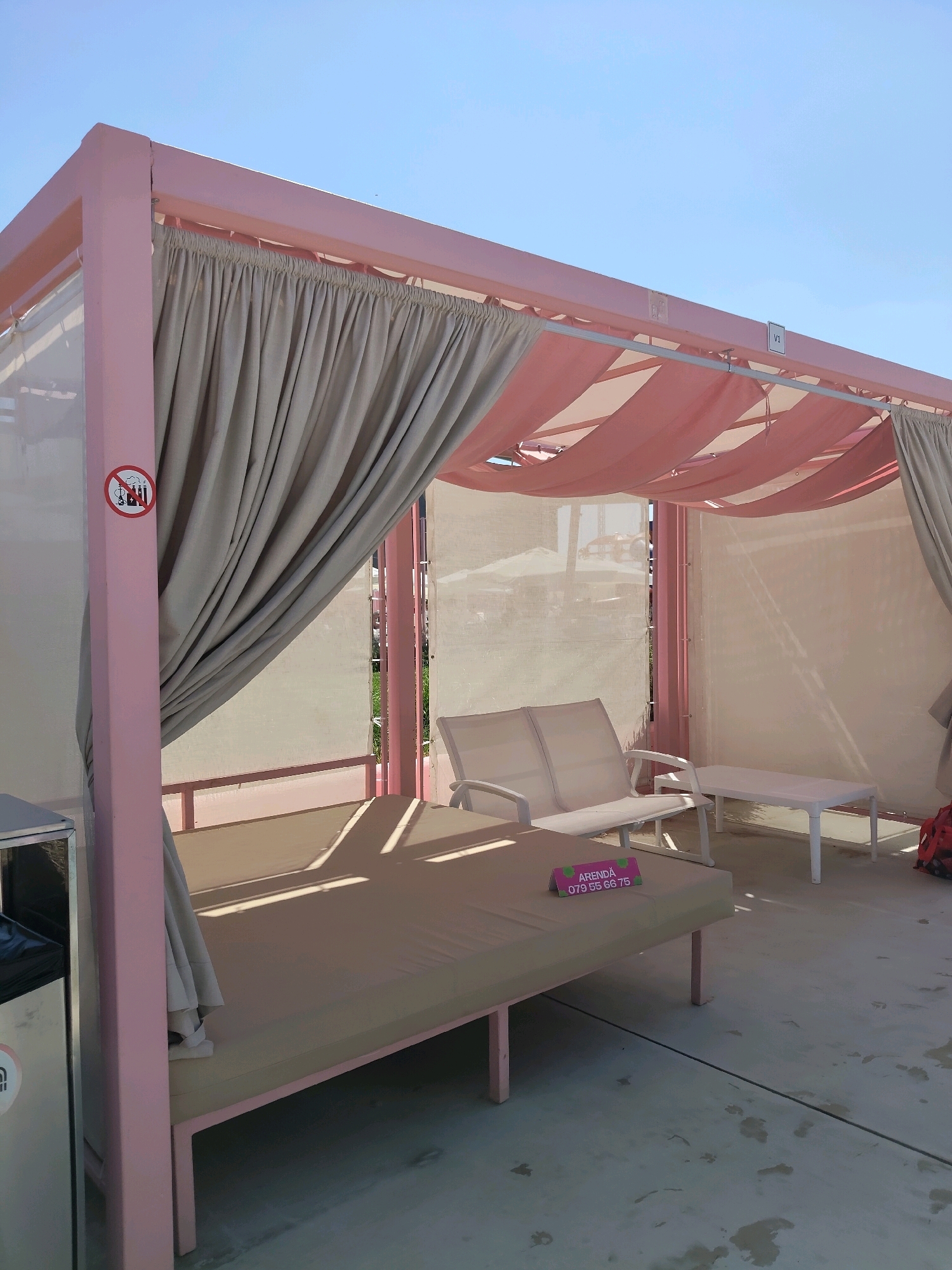 The image shows a pink outdoor cabana with beige curtains, located in Chișinău, Moldova. The cabana has a large cushioned daybed in the foreground and a two-seater bench and a small table in the background. A sign with the text "ARENDĂ 079 55 66 75" is placed on the daybed. A "no smoking" symbol is visible on the left support post of the cabana. The ground is paved with concrete, and there are shadows cast by the cabana structure. The sky is clear blue.