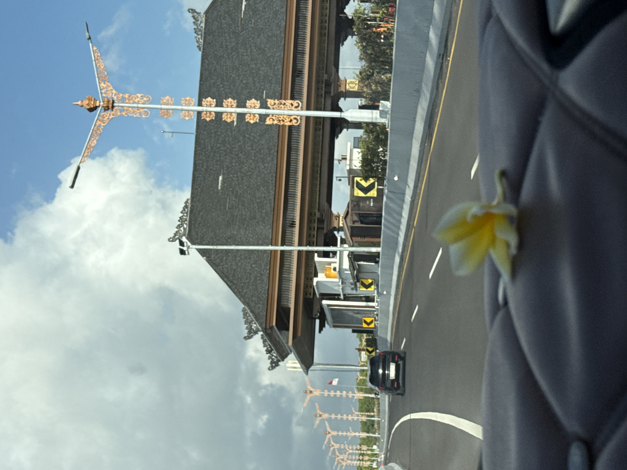 The image captures a view from inside a vehicle, looking forward towards a toll plaza in Kuta Selatan, Indonesia. The sky is partly cloudy with patches of blue. In the foreground, a yellow and white frangipani flower rests on a dark, quilted surface, possibly a dashboard. The road ahead is an asphalt highway with white lane markings and a yellow stripe on the right edge. A black car is traveling in the same direction. The toll plaza itself has a distinctive Balinese architectural style, featuring a dark, multi-tiered roof with ornate golden decorations on lampposts and building accents. Several lampposts with similar golden ornamentation line the side of the road leading up to the plaza. Traffic signs with yellow arrows indicating turns are visible. The environment suggests a warm, tropical climate.