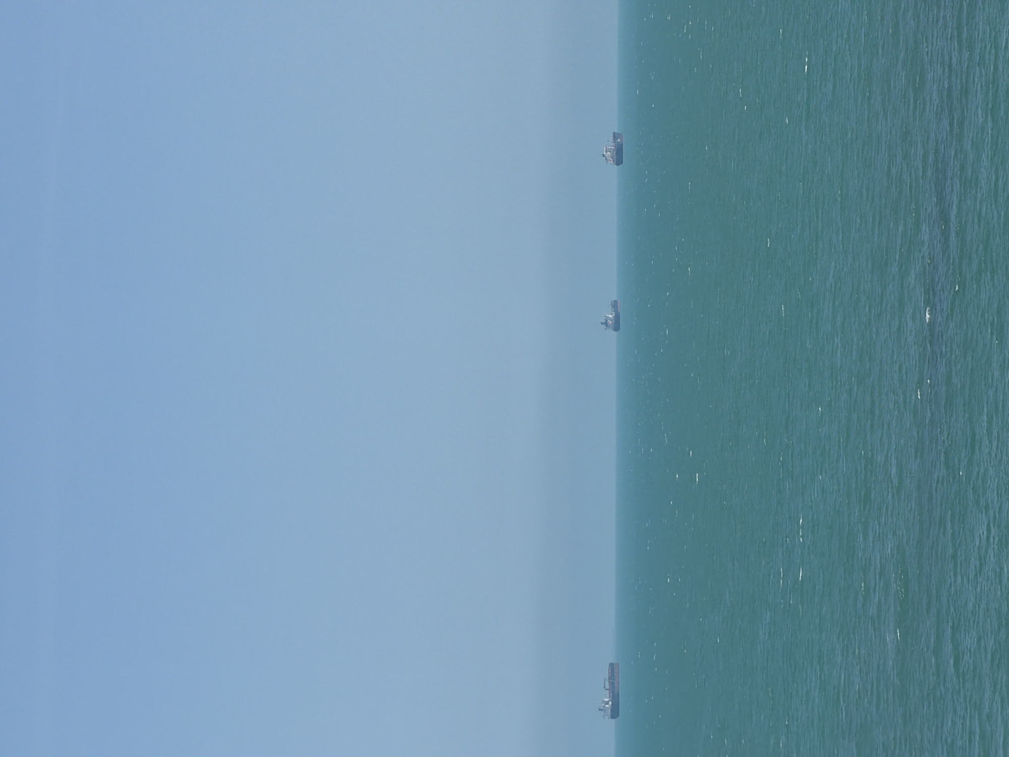 The image displays an expansive view of the open Black Sea off the coast of Burgas, Bulgaria, under a clear, bright sky. The upper two-thirds of the frame are dominated by an uninterrupted, light blue sky, devoid of clouds. A distinct but slightly hazy horizon line separates the sky from the sea. The lower third of the image features the sea, which presents a dynamic turquoise-to-dark-teal color. The water's surface is textured with numerous small ripples and scattered whitecaps, indicating active but not severe wave motion.
Three large cargo or bulk carrier vessels are visible on the distant horizon line, appearing as dark silhouettes against the brighter sky and sea. They are positioned horizontally across the frame, seemingly static or moving very slowly. From left to right:
1. The leftmost vessel is the largest and most defined, with a dark hull and a lighter-colored superstructure towards the stern.
2. The central vessel is the smallest and most indistinct, appearing further away and partially obscured by atmospheric haze.
3. The rightmost vessel is slightly larger than the central one but smaller than the leftmost, also exhibiting a dark hull and faint superstructure details.
No people are visible in the scene. There are no other objects or structures, emphasizing the vastness of the sea and sky. The overall environment is one of open maritime space, characterized by clear weather conditions.
