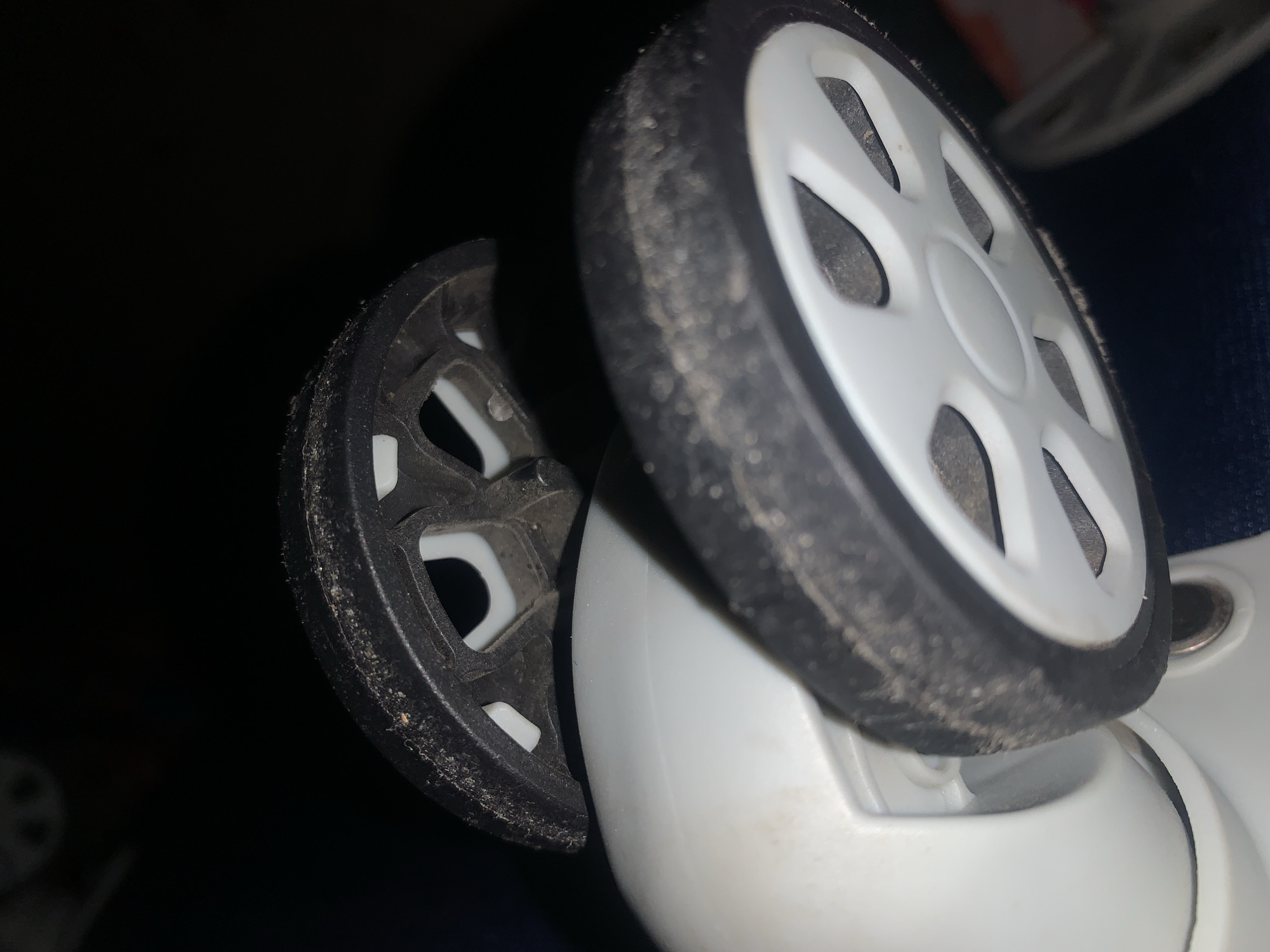 The image displays a close-up, low-angle shot of two wheels, likely from a suitcase or trolley. Both wheels are black rubber with white plastic spokes and appear somewhat dirty. The foreground wheel is larger and more in focus, showing a textured rubber surface with visible dust and debris. The background wheel is smaller, darker, and out of focus, positioned above and behind the foreground wheel. The wheels are attached to a white plastic casing. The environment is dark, with the exception of the illuminated wheels and casing. There is no discernible urban environment or specific location information within the image that would allow for pinpointing it to Jalingo, Nigeria.
