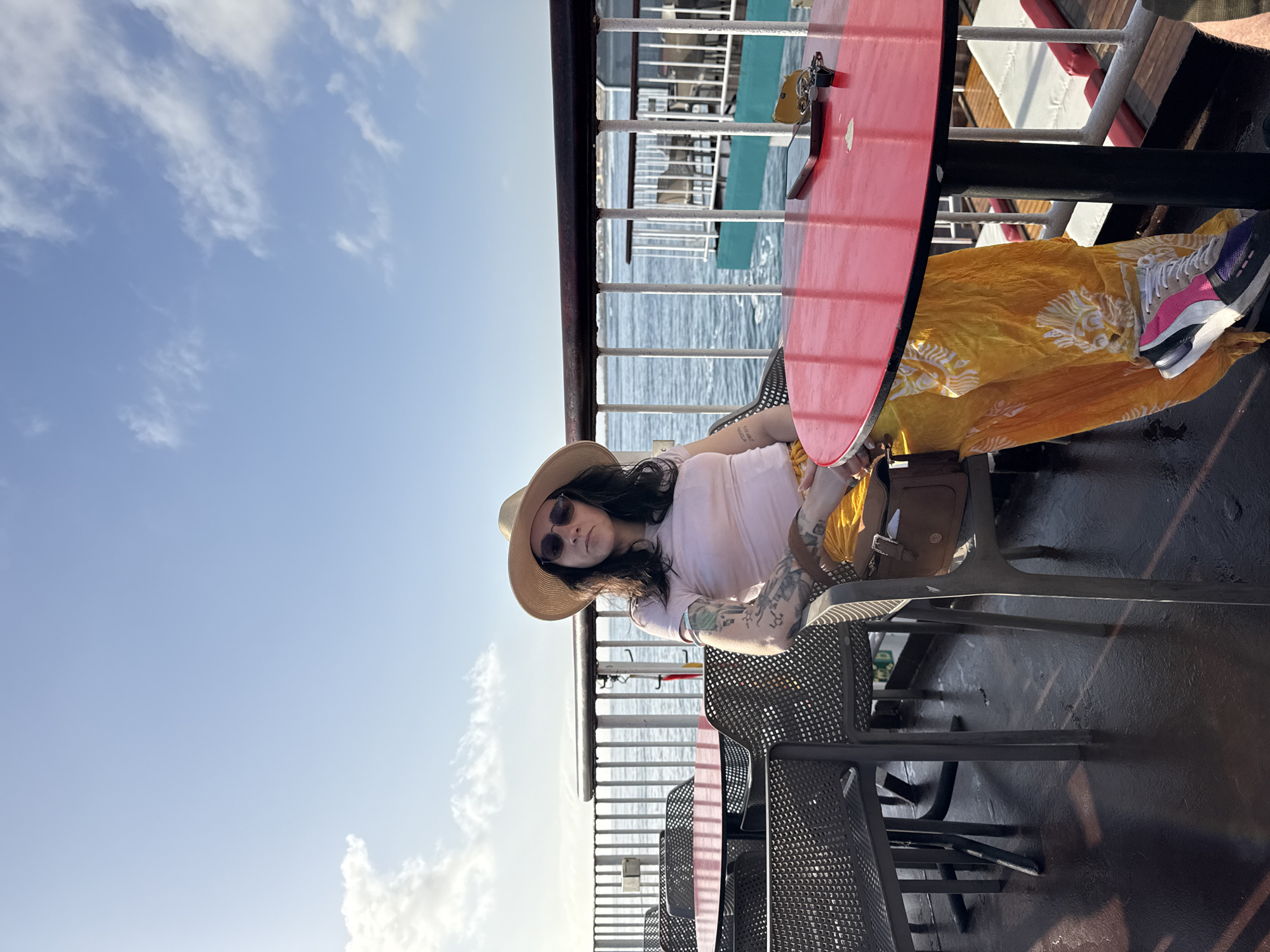A woman sits at a red table on a ferry or boat under a blue sky with clouds. She wears a light pink top, a straw hat, and sunglasses. Her left arm has tattoos. A yellow patterned cloth rests on her lap. She is wearing white, pink, and grey sneakers. A brown purse sits next to her on the chair. The ferry has metal railings and perforated metal chairs. The water can be seen beyond the railings. The location is likely Corralejo, Spain based on the surroundings.