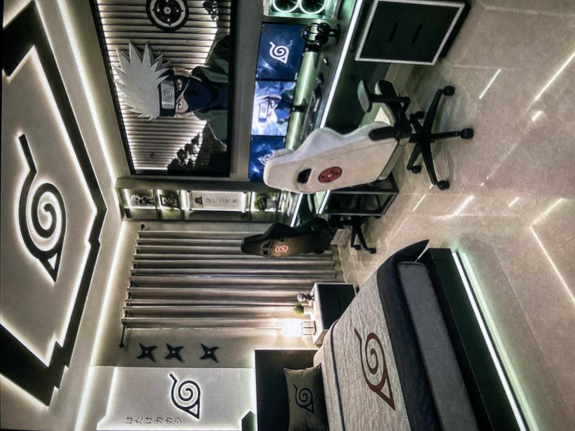 The image depicts a bedroom interior with a prominent anime theme. The walls are decorated with images and symbols related to the "Naruto" series, including characters like Kakashi and the Hidden Leaf Village symbol. A bed is present with bedding also displaying the Hidden Leaf Village symbol. There are black ninja star decals on the wall. A computer desk area features multiple monitors and a gaming chair. The room is illuminated with ambient lighting, including lighted border features, creating a modern, stylized aesthetic. The flooring appears to be tiled. There is no information available within the image confirming a specific location in the city of Lhokseumawe, Indonesia.