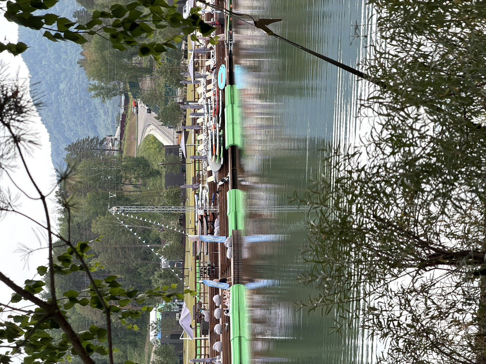 This is an outdoor scene at a lake with a wooden pier. Several people are visible on the pier, some sitting in chairs, others standing. There are various objects on the pier, including lounge chairs, umbrellas, and paddleboards or kayaks lined up. Green safety barriers are positioned along the edge of the pier. In the background, there are trees and a forested hillside, with a road and some vehicles visible on it. A tall white metal structure with triangular flags is also present, suggesting an event or competition might be taking place. The water reflects the sky and surrounding elements, indicating it is likely daytime, and the overcast sky suggests cloudy or mild weather. The overall impression is of a recreational area by a lake, possibly during a public gathering or a sporting event. Text visible on blue banners on the pier appears to read "ЦЕНТР ПЕДАГОГИЧЕСКОЙ АКТИВНОСТИ".