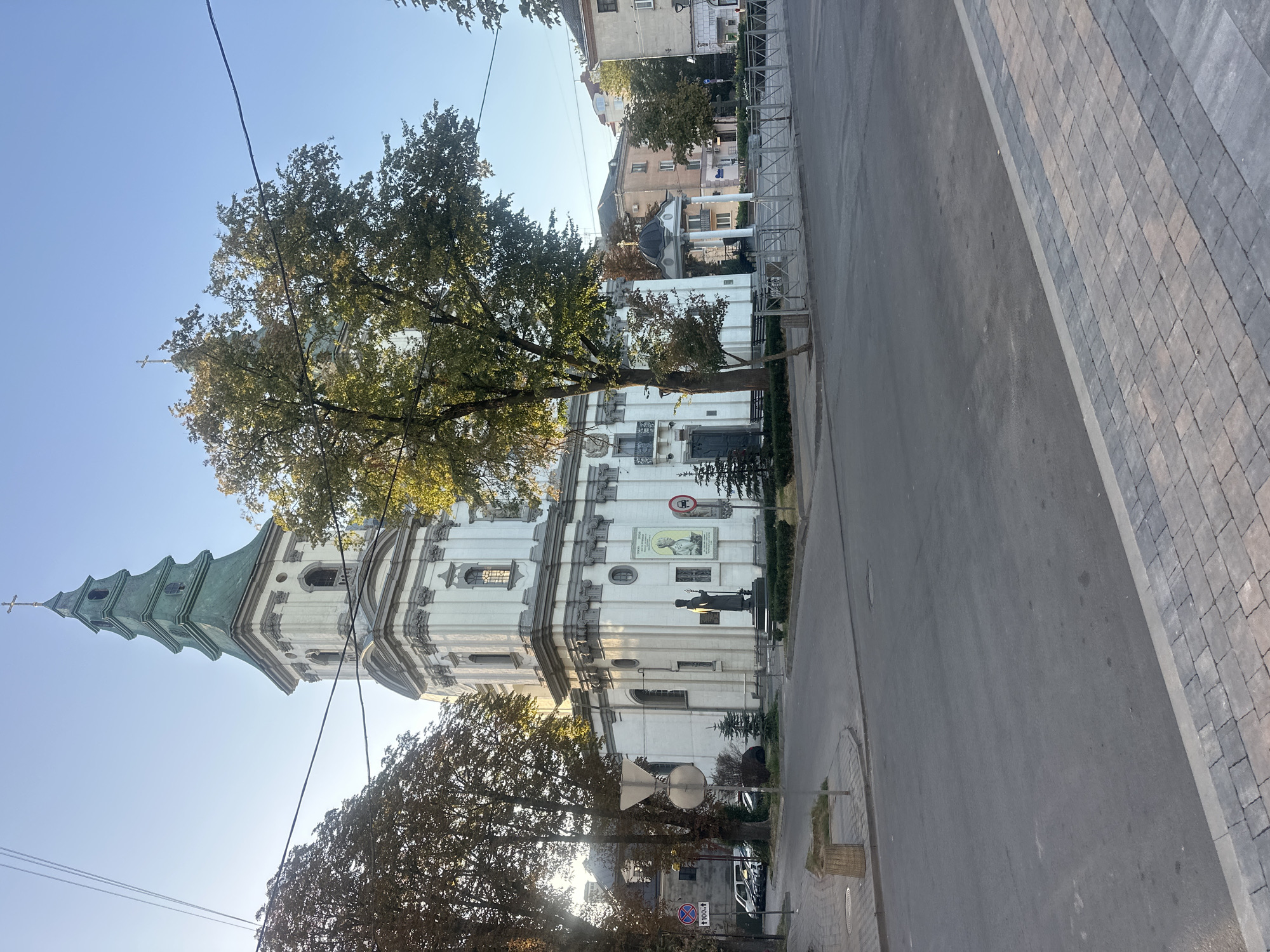 The image shows the Church of the Immaculate Conception of the Blessed Virgin Mary in Ternopil, Ukraine. The church features a tall, multi-tiered green spire topped with a golden cross. The main facade of the church is white with gray architectural details, including columns and decorative carvings. A statue of a figure in religious attire stands in front of the church. The church is situated on a street corner, with a paved road and sidewalk in the foreground. Trees with autumnal foliage are visible on either side of the church. In the background, other buildings and utility wires can be seen. The sky is clear and blue, indicating daytime.