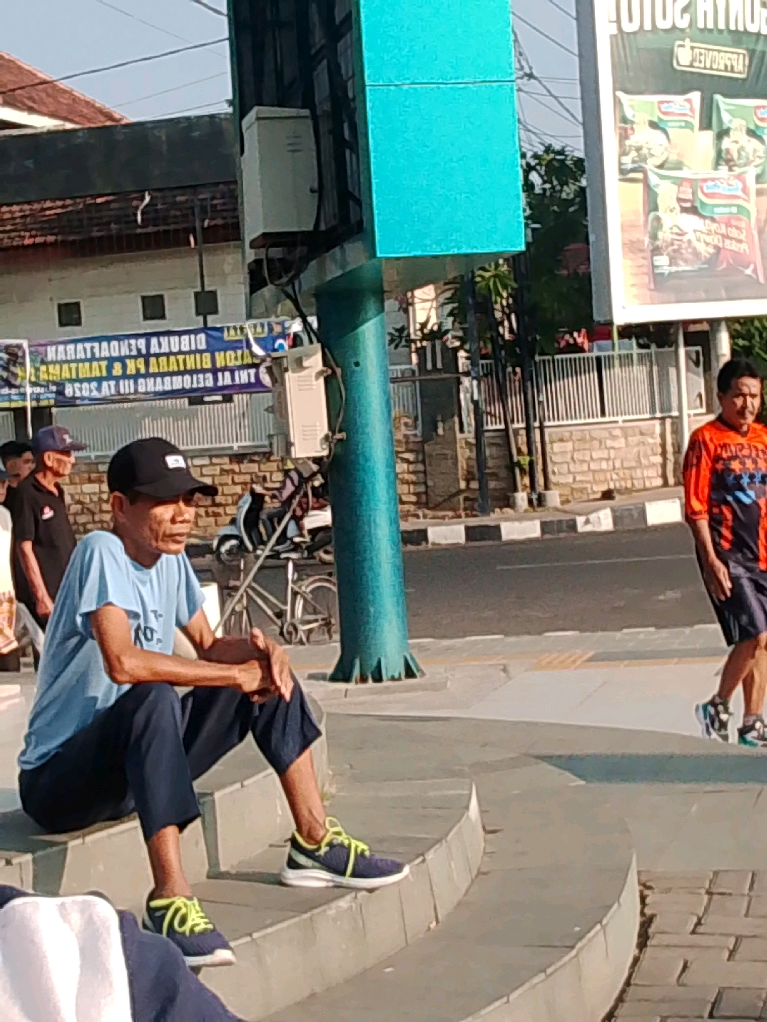 This image depicts a street scene in Probolinggo, Indonesia, characterized by its urban environment and moderate daylight. The primary focus is on a man seated on concrete steps, wearing a blue t-shirt and dark pants. He is looking to his left, with his hands clasped. To his right, another man in an orange and black athletic shirt and dark shorts walks away from the camera. In the background, a billboard advertising Indomie noodles is prominently displayed. A bicycle leans against a wall near the seated man. The presence of a traffic light pole and the general street layout suggest this is a public area within the city. The overall scene is that of everyday life in a bustling Indonesian city.