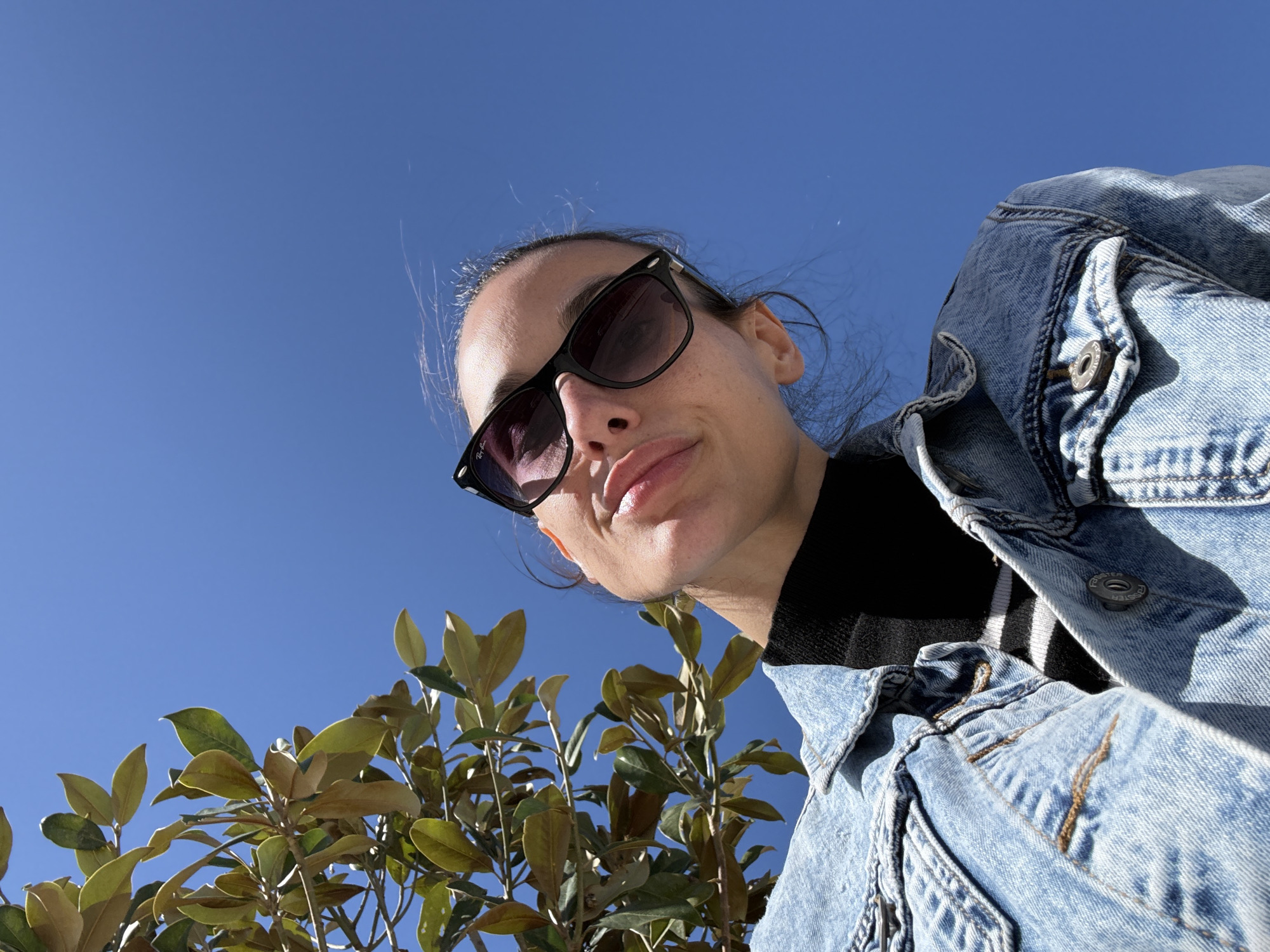 A low-angle shot depicts a woman wearing sunglasses and a denim jacket against a clear blue sky. Green foliage is visible in the lower-left corner. The photo was not taken in Mouzouras, Greece; it appears to be taken at an urban location with a tree.