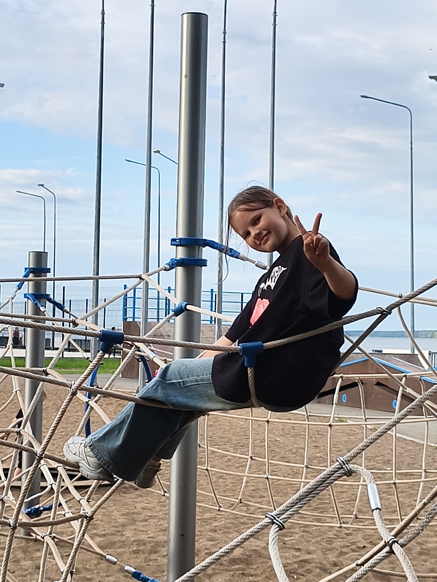 A young girl with dark hair and a playful expression is perched on a rope climbing structure at an outdoor playground. She is wearing a black t-shirt with a graphic design and blue jeans, along with light-colored sneakers. Her right hand is raised, forming a peace sign. The playground features thick ropes forming a net-like structure, connected to metal poles with blue fasteners. The ground beneath is covered in sand. In the background, there are more playground elements, fences, lampposts, and a body of water, suggesting a location near a river or lake. The sky is partly cloudy, indicating it is daytime. The overall scene is cheerful and suggests a child enjoying a day at the playground.